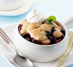 Close-up of blueberry cobbler in a white ramekin topped with whipped cream and a mint leaf, spoon at the side on a light plate