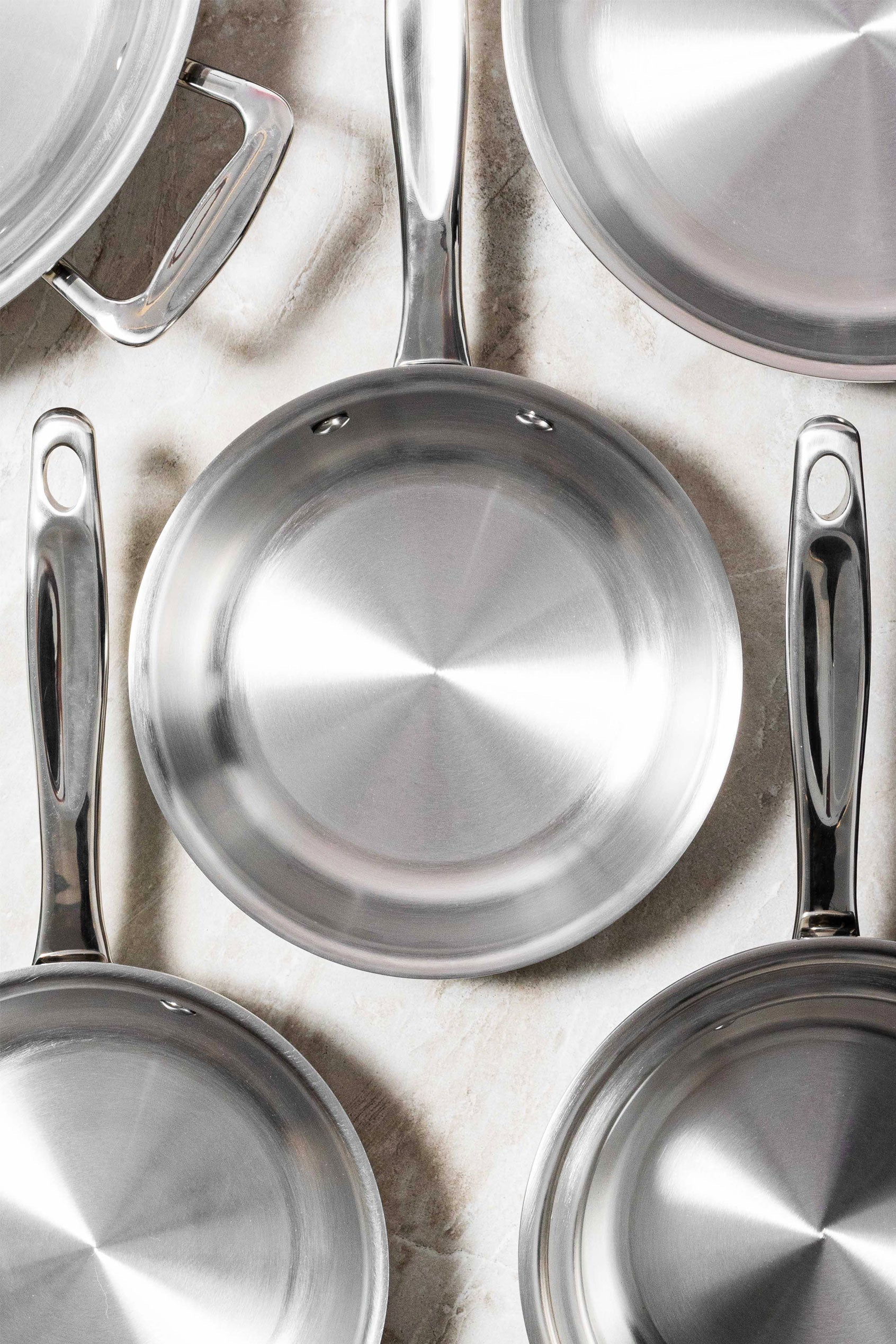 Top-down view of stainless steel frying pans showcasing polished interiors and riveted handles on a light stone surface