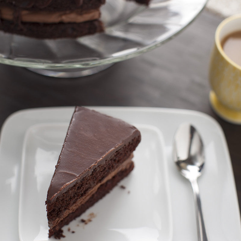 A slice of double chocolate skillet cake on a white plate with a spoon and a cup of coffee in the background.