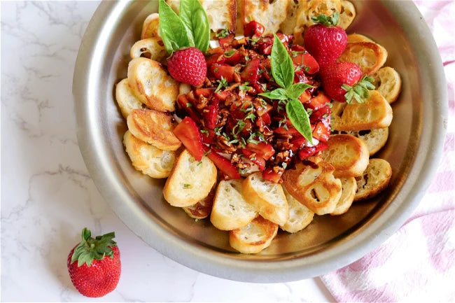 A platter of toasted baguette slices topped with balsamic strawberries and fresh basil, garnished with whole strawberries.