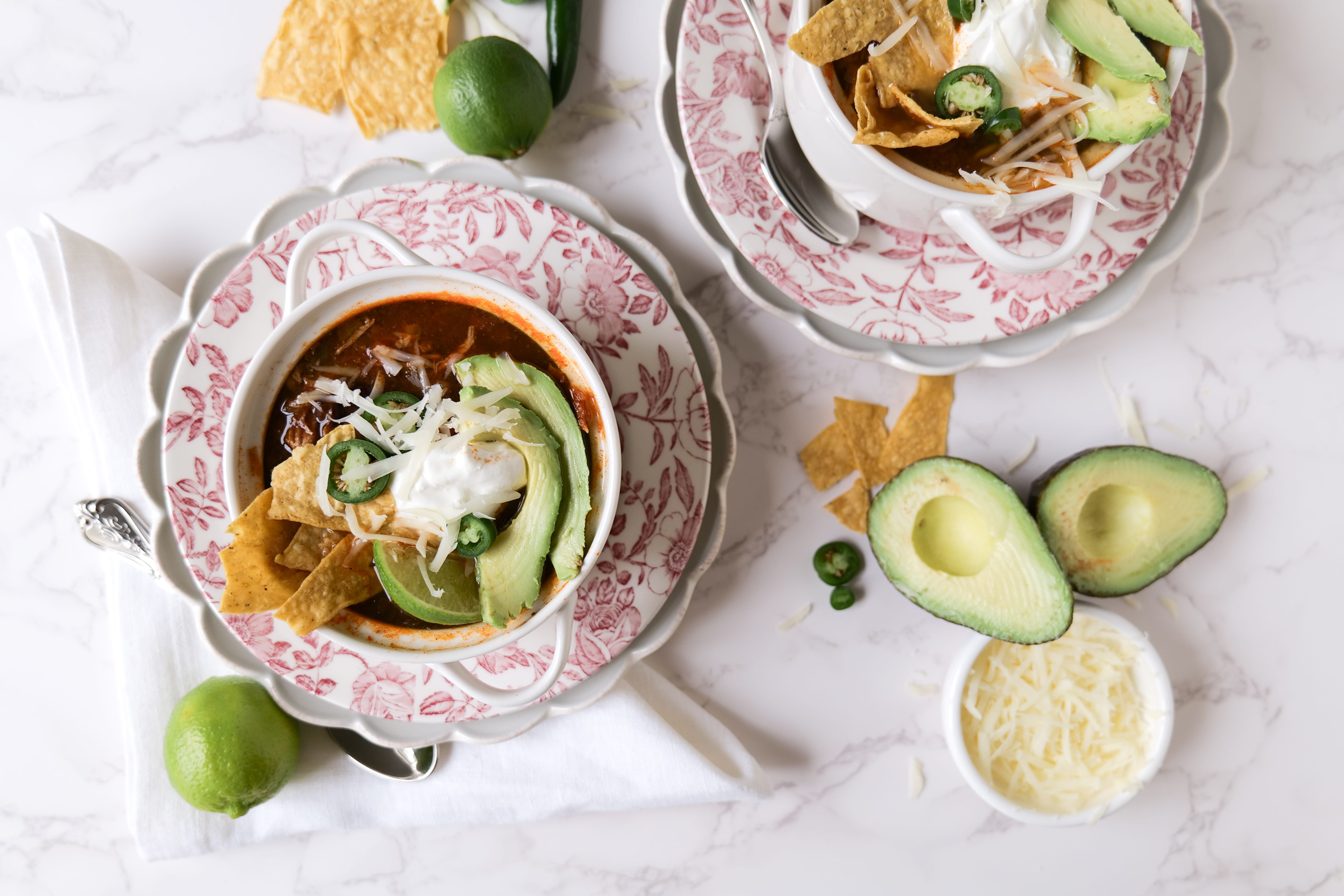 Bowl of Chicken Tortilla Soup topped with avocado, jalapenos, tortilla chips, and sour cream, served with lime and cheese on the side.