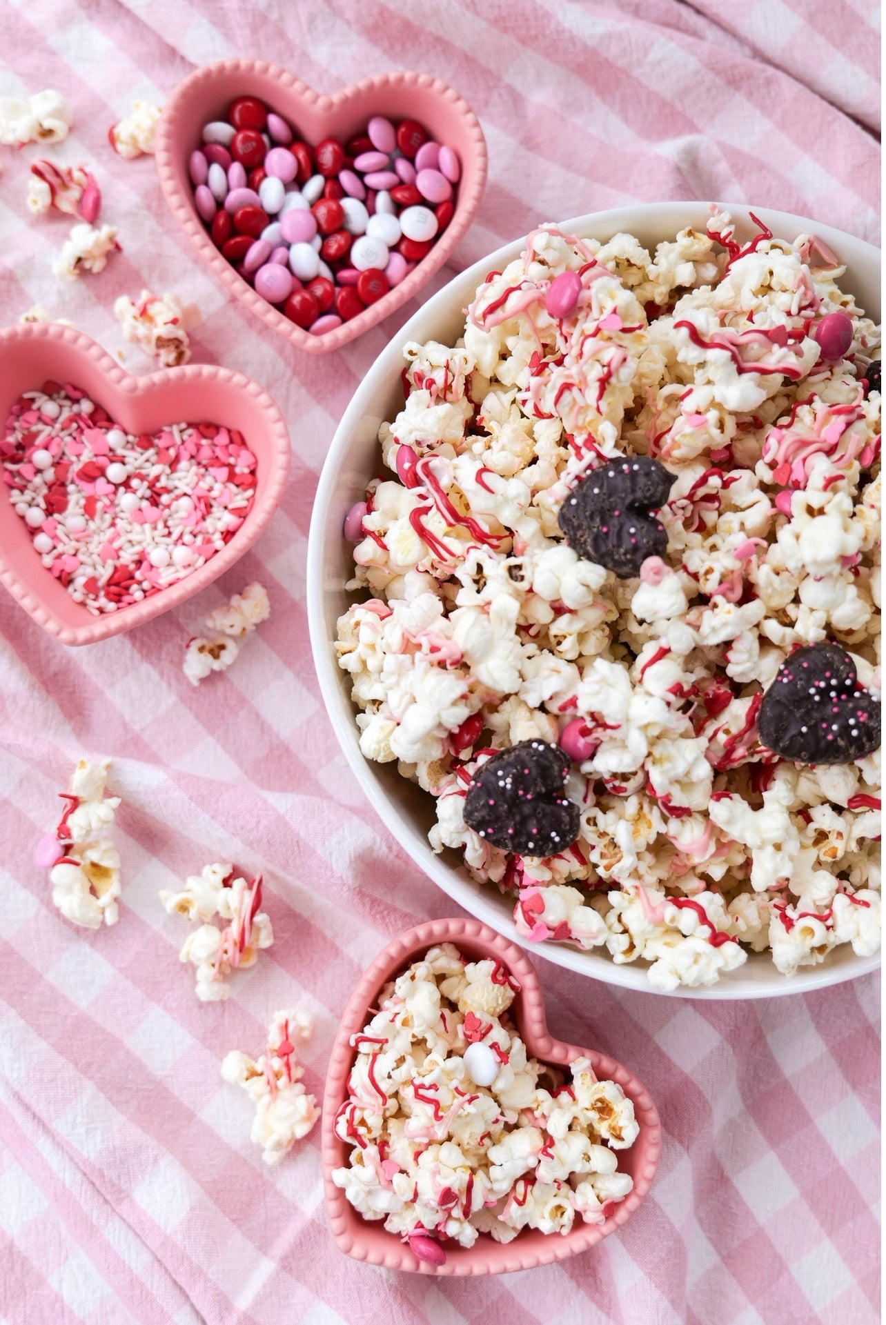 Bowl of white chocolate popcorn drizzled with pink and red chocolate, surrounded by heart-shaped bowls of candy and sprinkles.