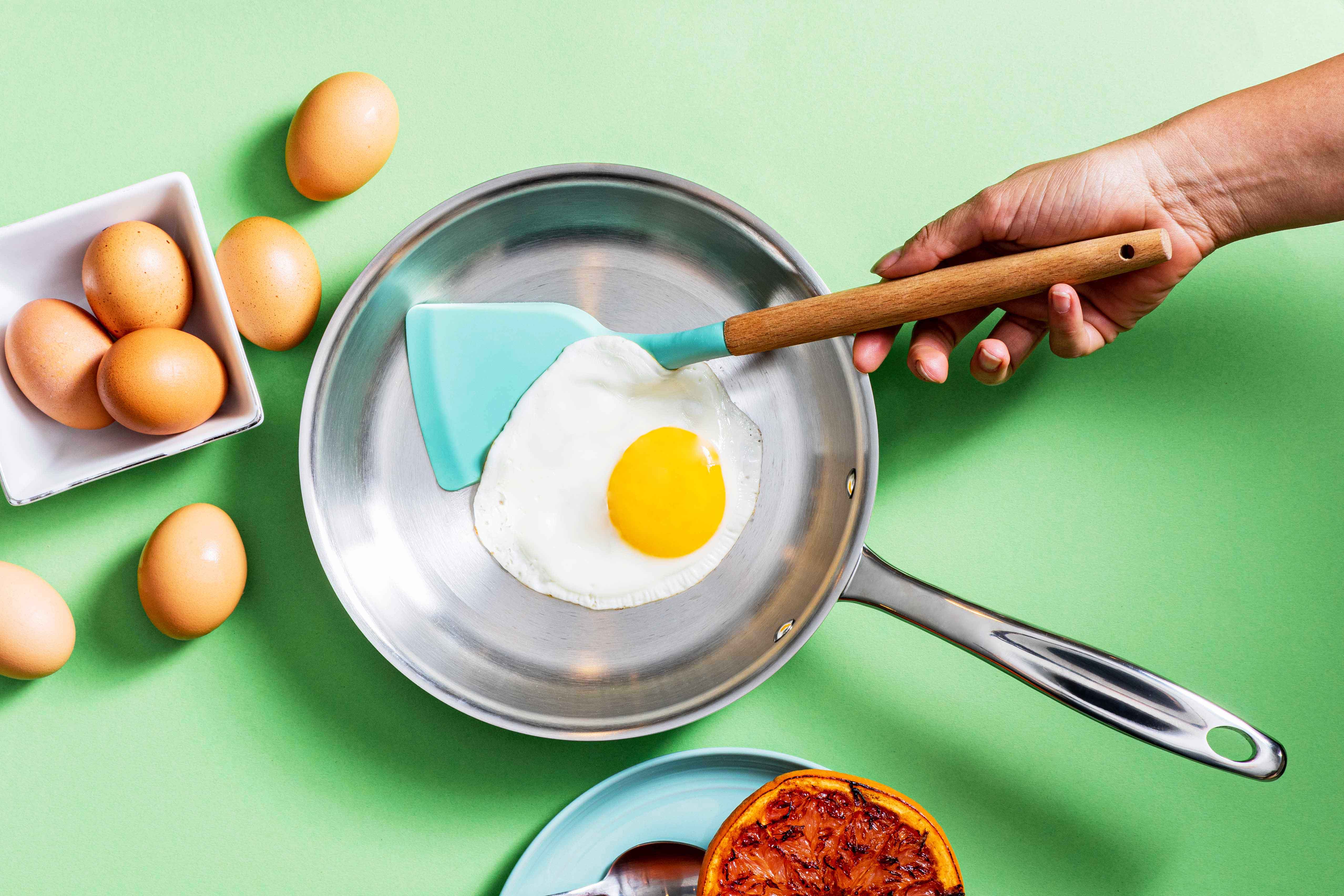 A hand using a spatula to flip a fried egg in a stainless steel pan, with raw eggs nearby.