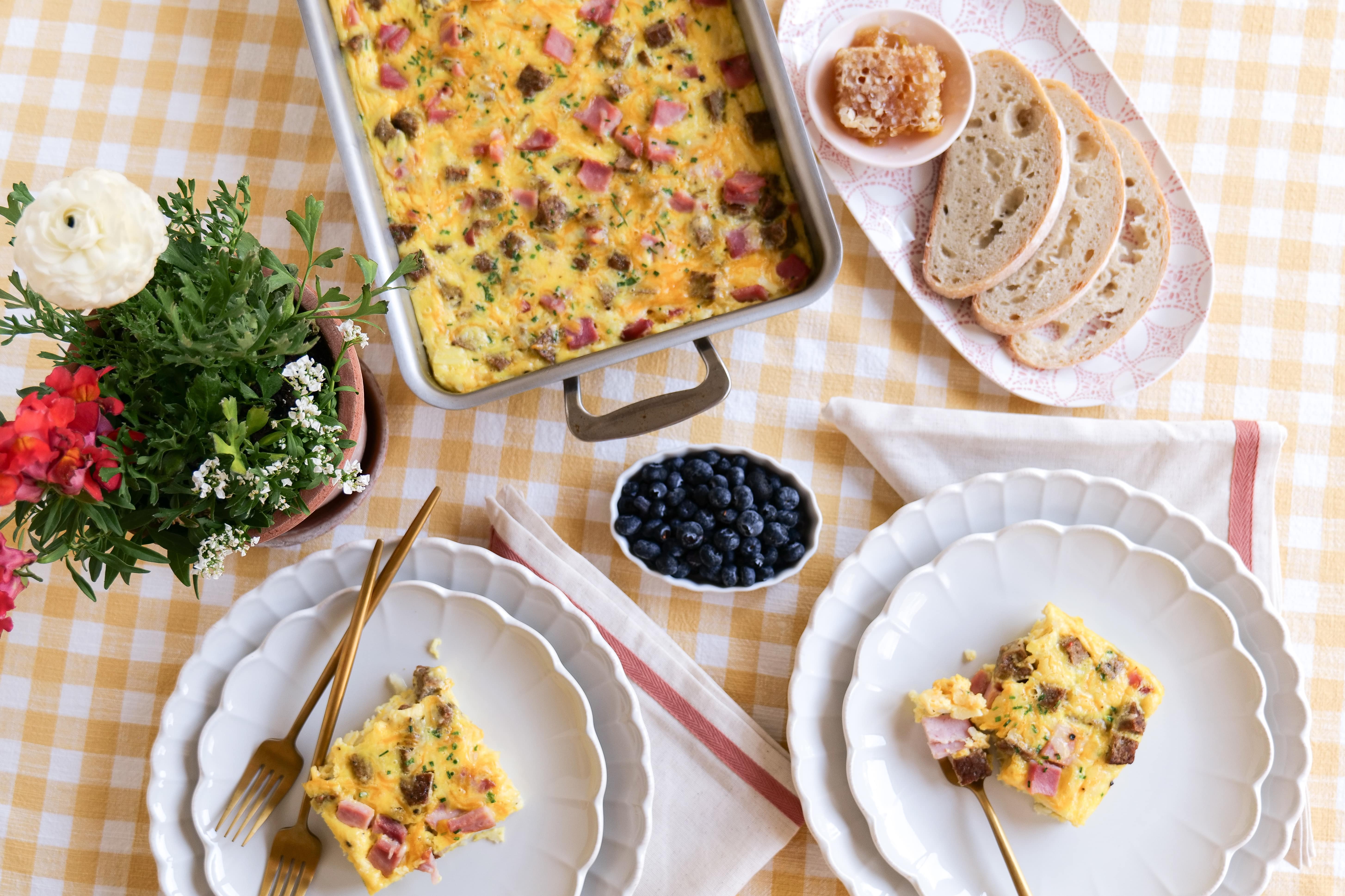 A serving of breakfast casserole with ham and sausage on a table with bread and blueberries