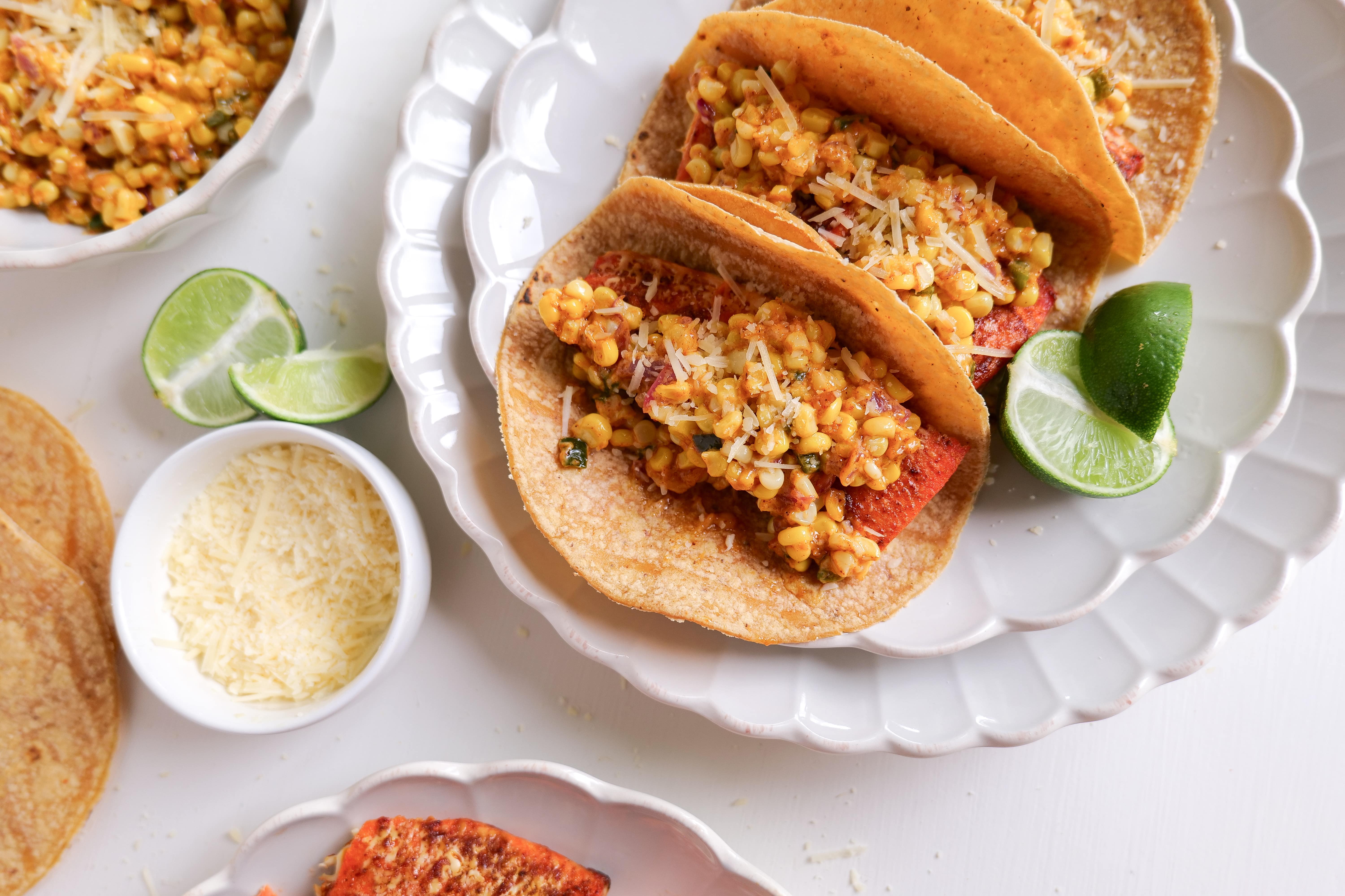 Three salmon tacos topped with elote salsa, served with lime wedges and cheese on the side.