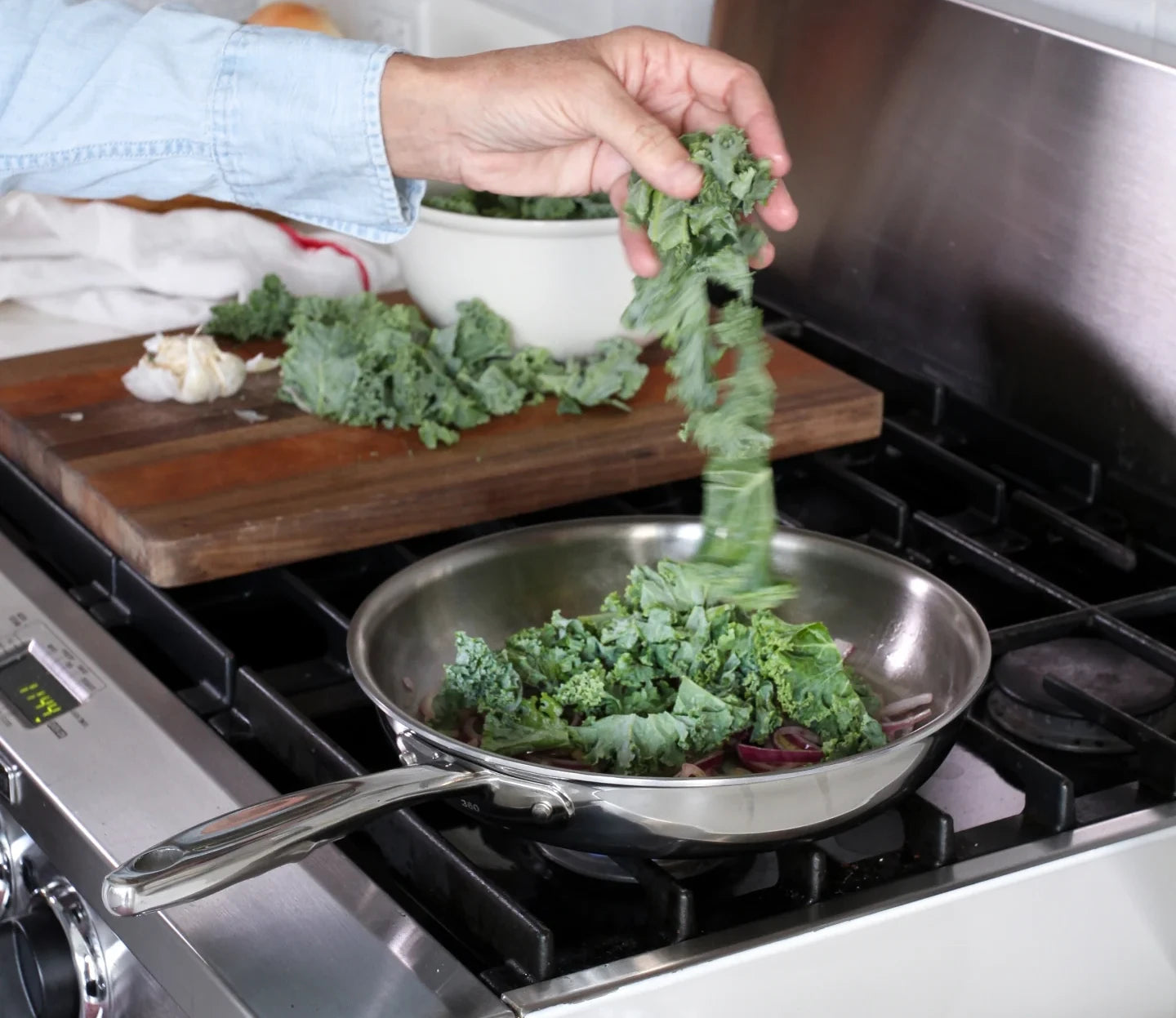 A hand adding kale to a stainless steel frying pan on a stove with chopped garlic and a bowl of kale in the background.