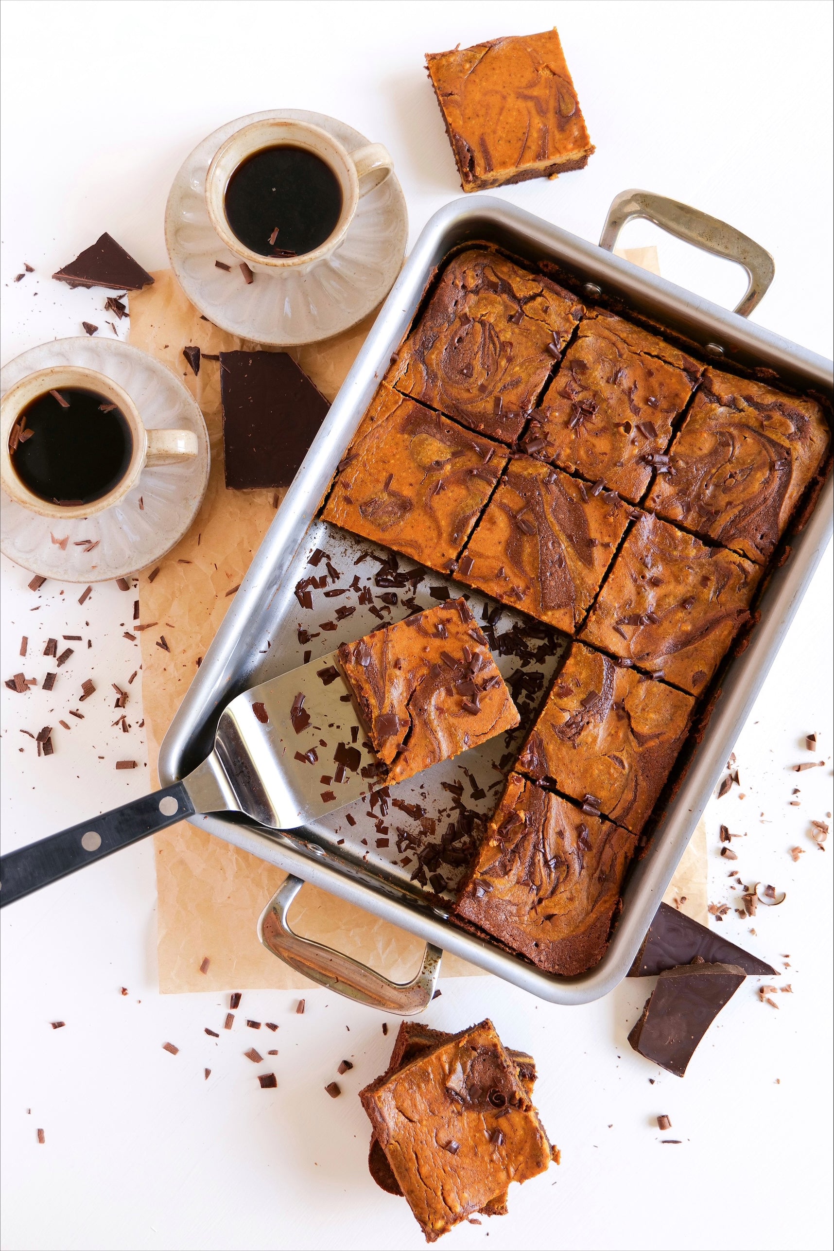 Top-down pumpkin swirl brownies cut in a metal baking pan with two coffee cups and chocolate shards on a white background