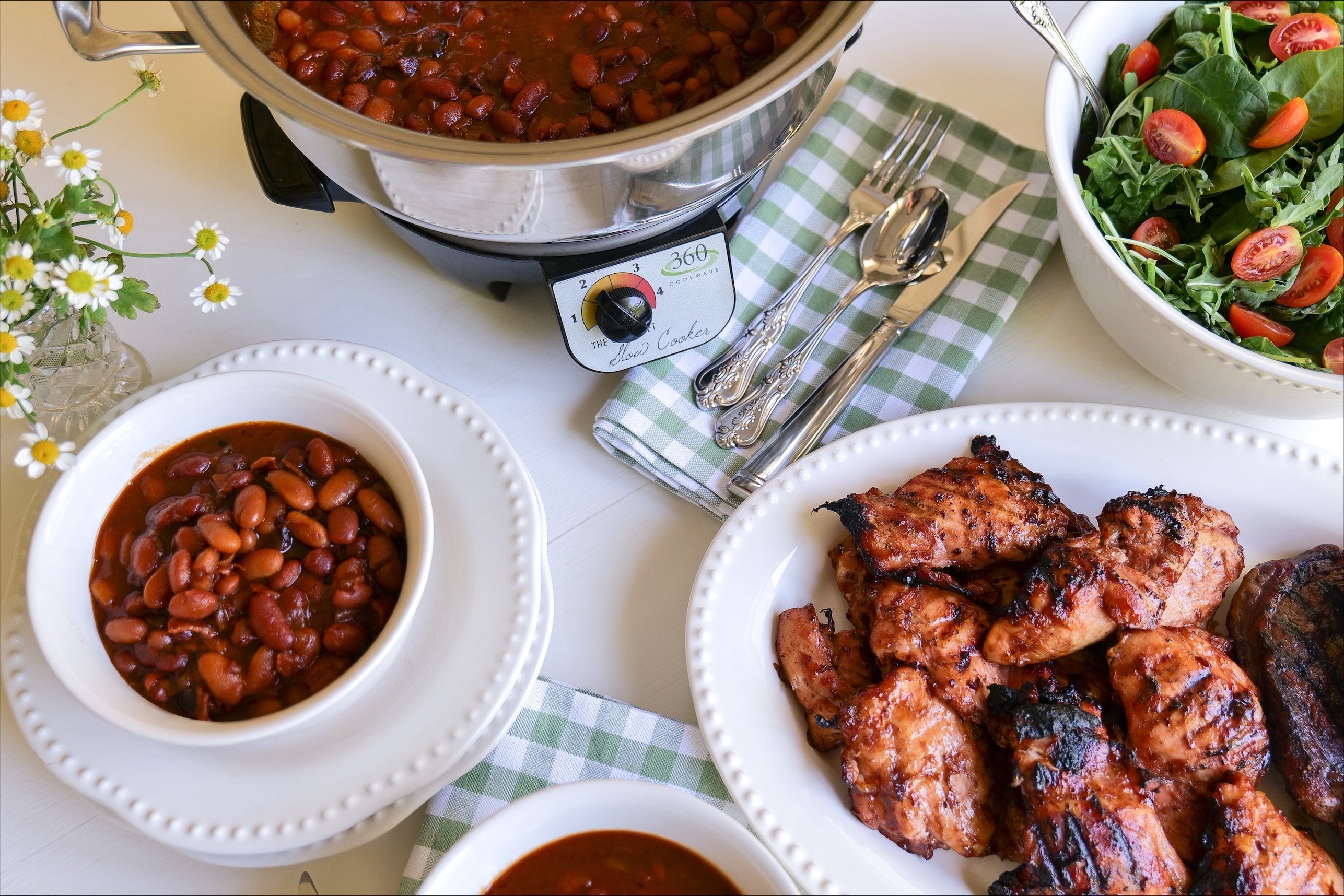 Slow cooker full of baked beans with bowls of beans, grilled chicken and a tomato salad on a table with a green checkered napkin