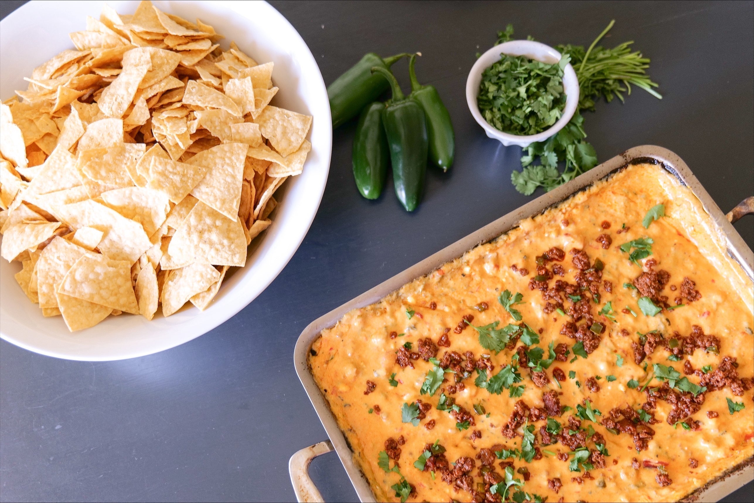 Bowl of tortilla chips beside fresh jalapeños and a pan of cheesy chorizo queso topped with cilantro on a dark tabletop