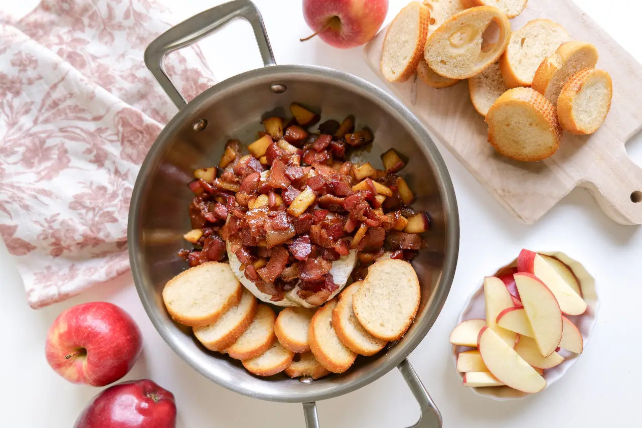 Baked brie topped with bacon and diced apples in a stainless steel pan, served with sliced baguette and apple wedges