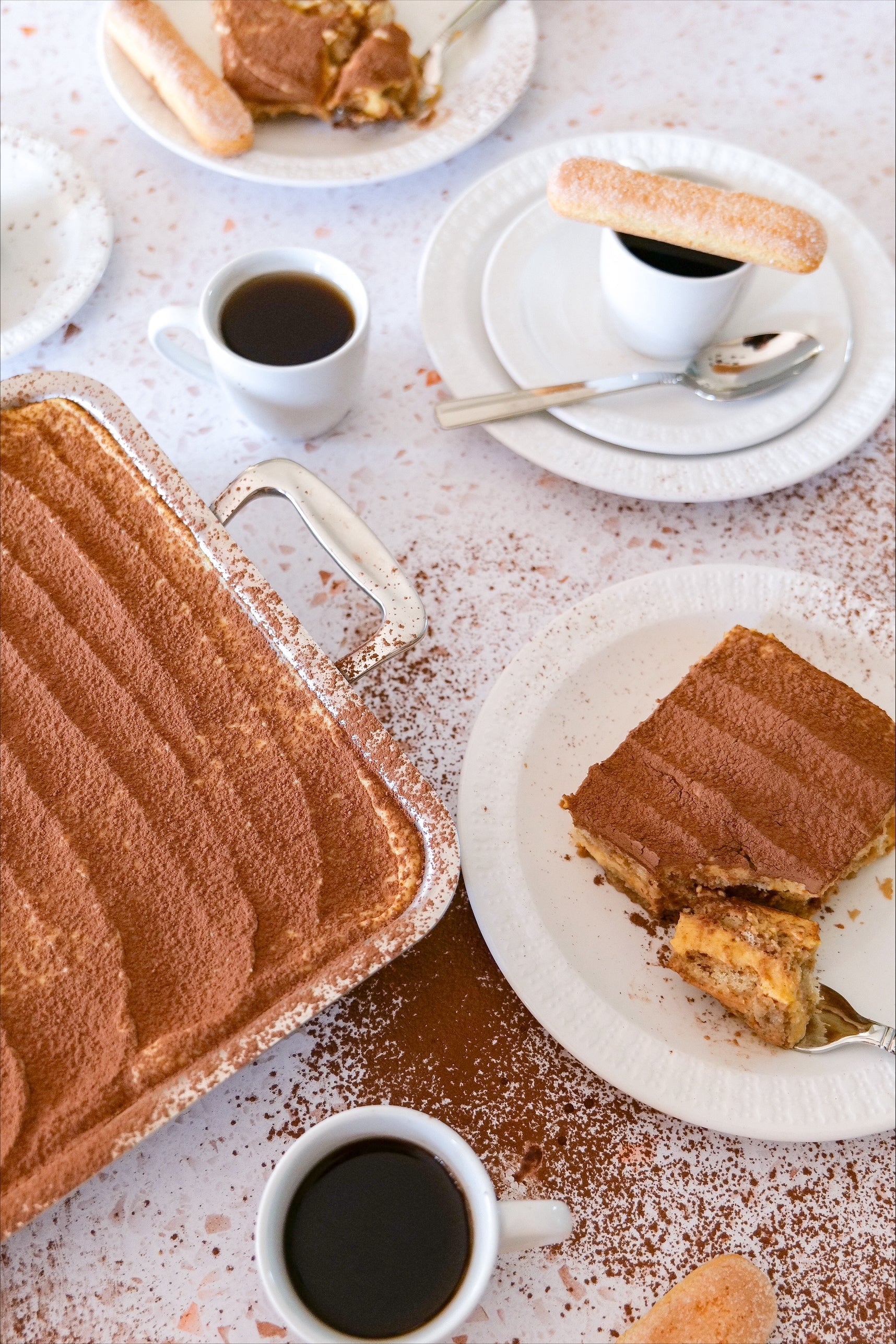 Pumpkin tiramisu served in a baking dish with cocoa-dusted plated slices, espresso cups, and ladyfingers on a speckled table