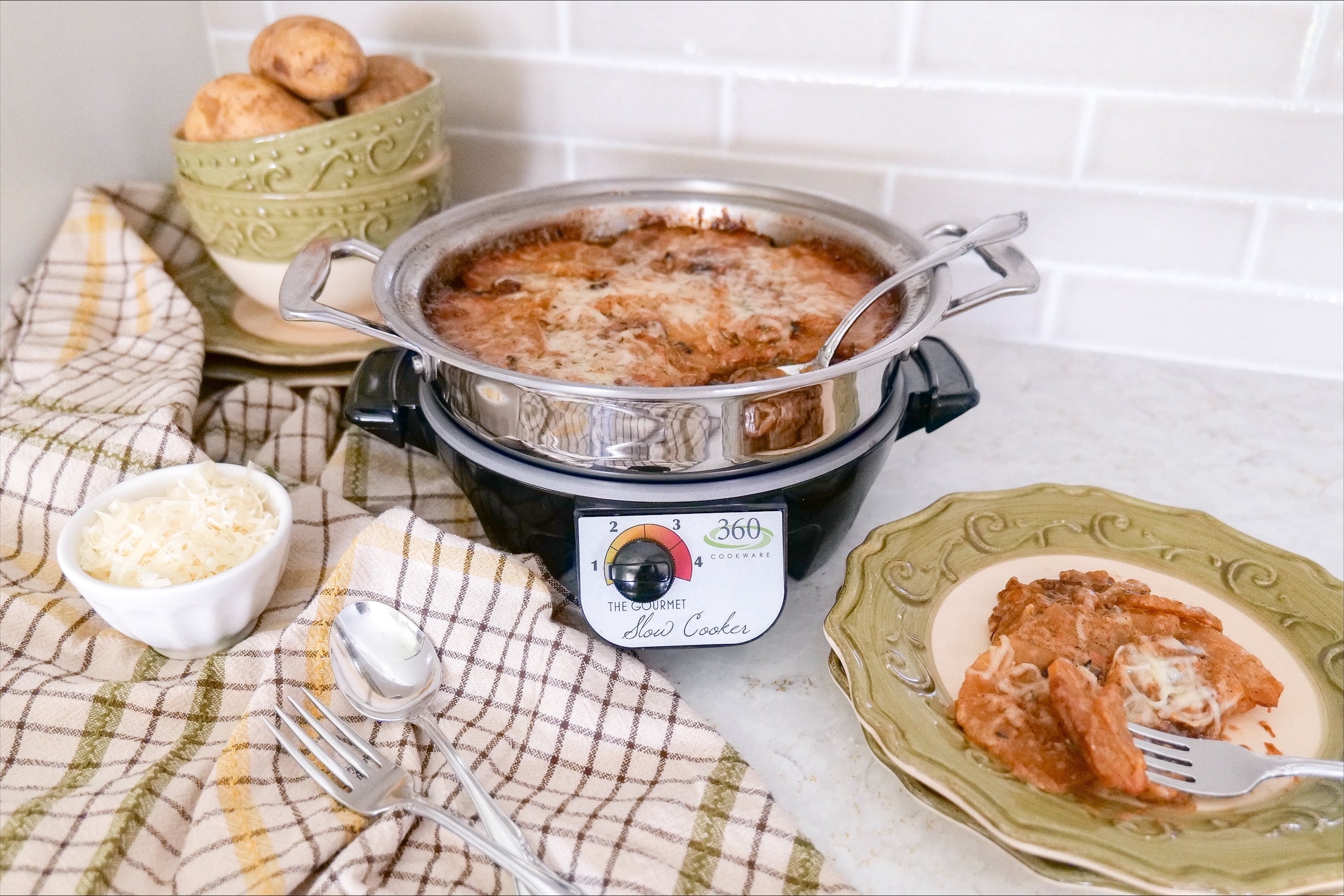 Slow cooker with bubbling cheesy tomato casserole on a kitchen countertop with bowls of shredded cheese and potatoes