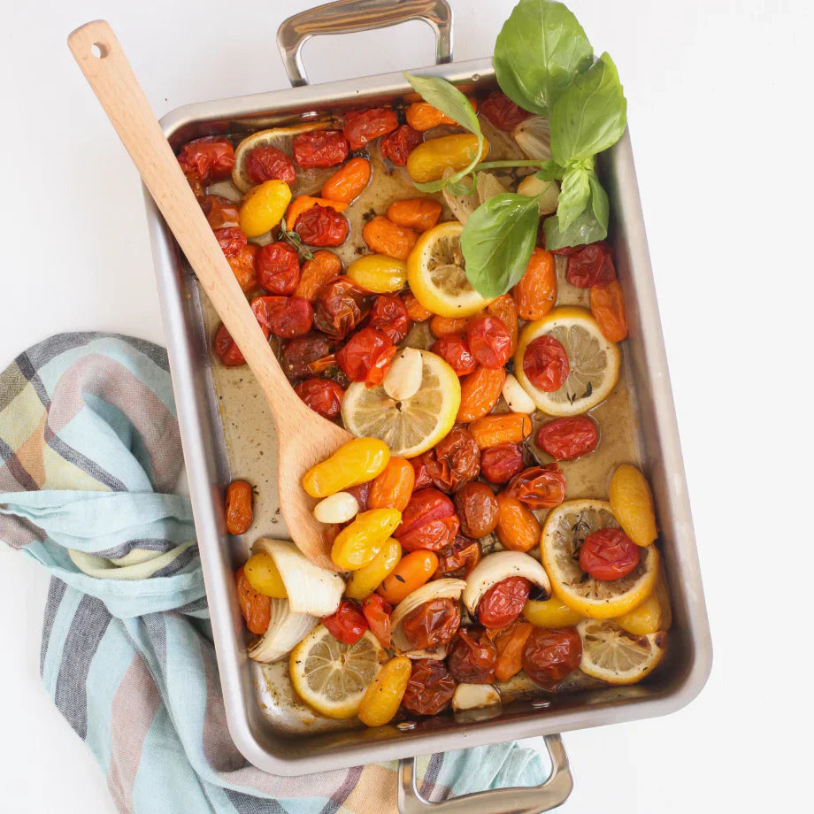 Overhead view of rectangular stainless steel roasting pan with handles showcasing roasted cherry tomatoes, lemon slices, garlic and basil on a striped tea towel