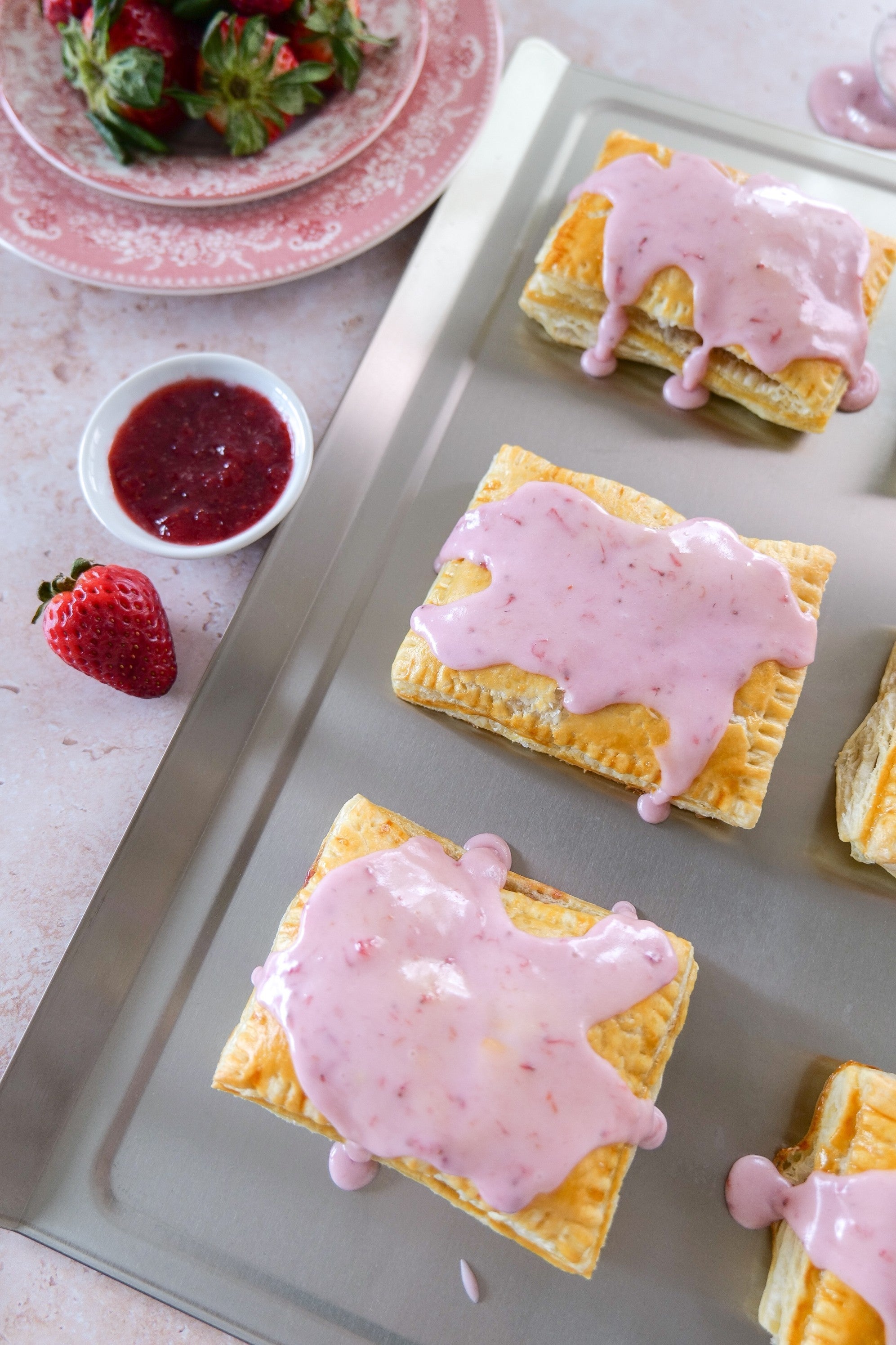 Strawberry-glazed puff pastry turnovers arranged on a baking sheet with fresh strawberries and a small bowl of strawberry jam