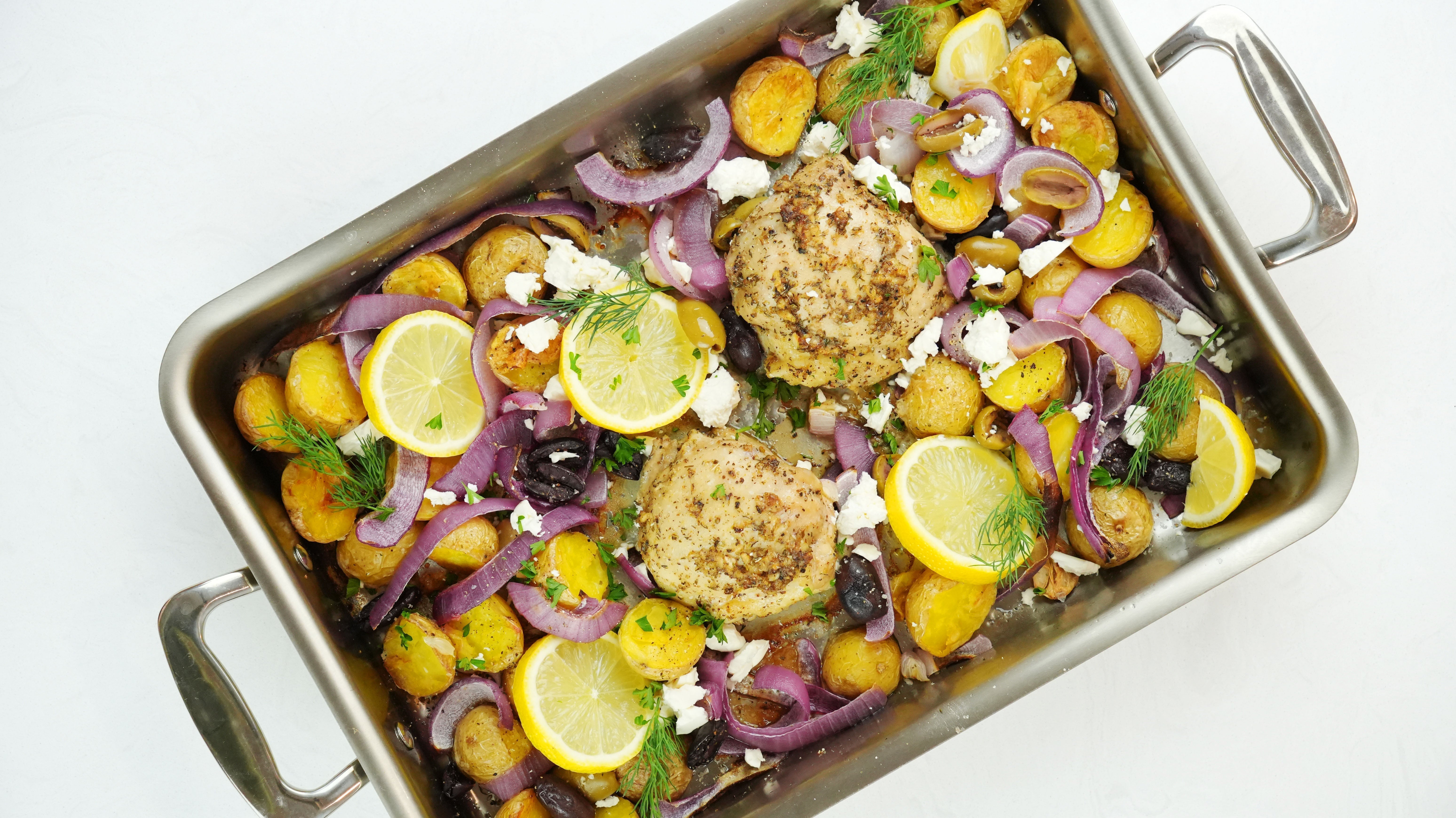 Roasted chicken thighs with baby potatoes, red onion, lemon slices, olives and feta in a stainless steel roasting pan