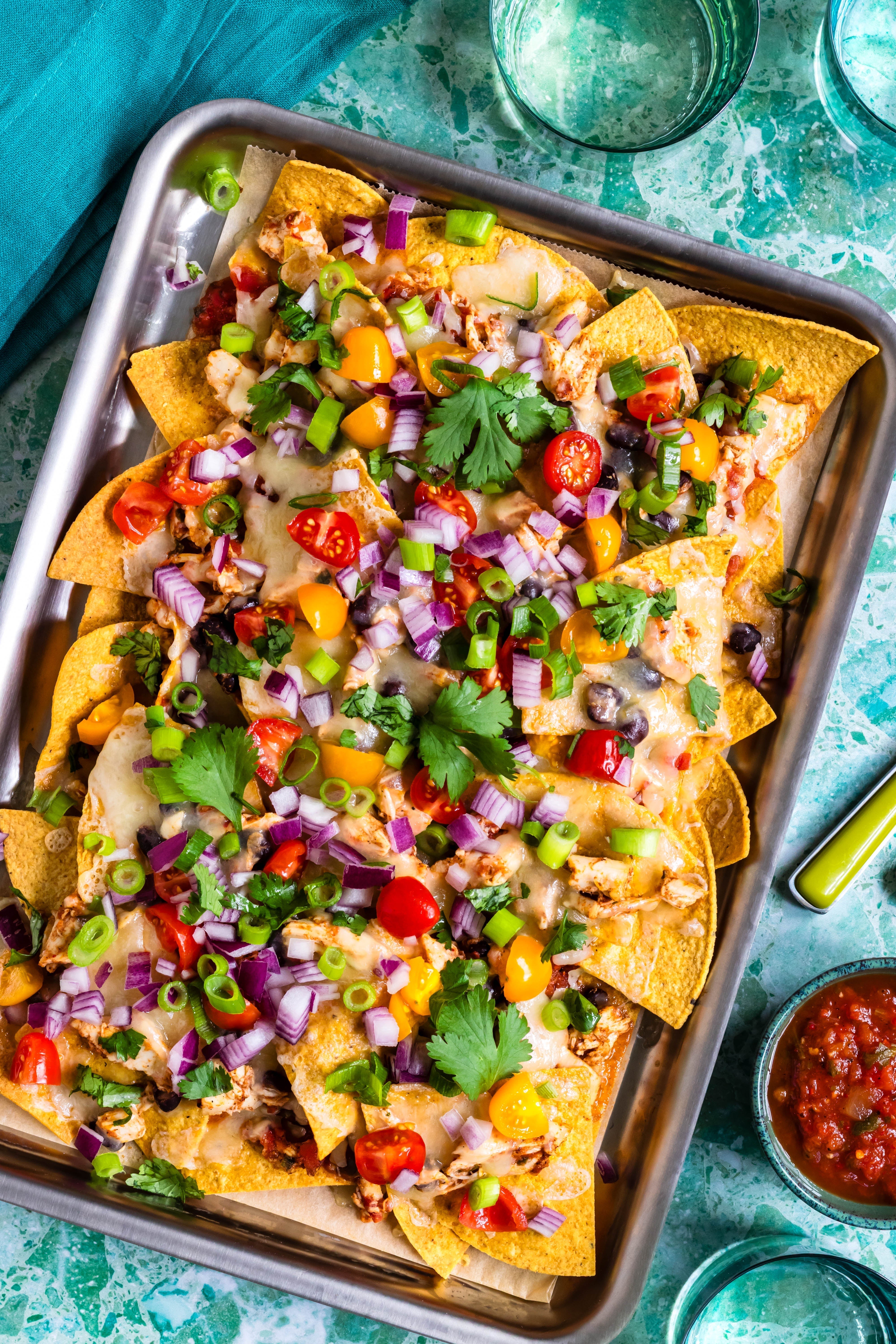 Sheet-pan nachos with melted cheese, black beans, cherry tomatoes, diced red onion and cilantro on a metal baking sheet