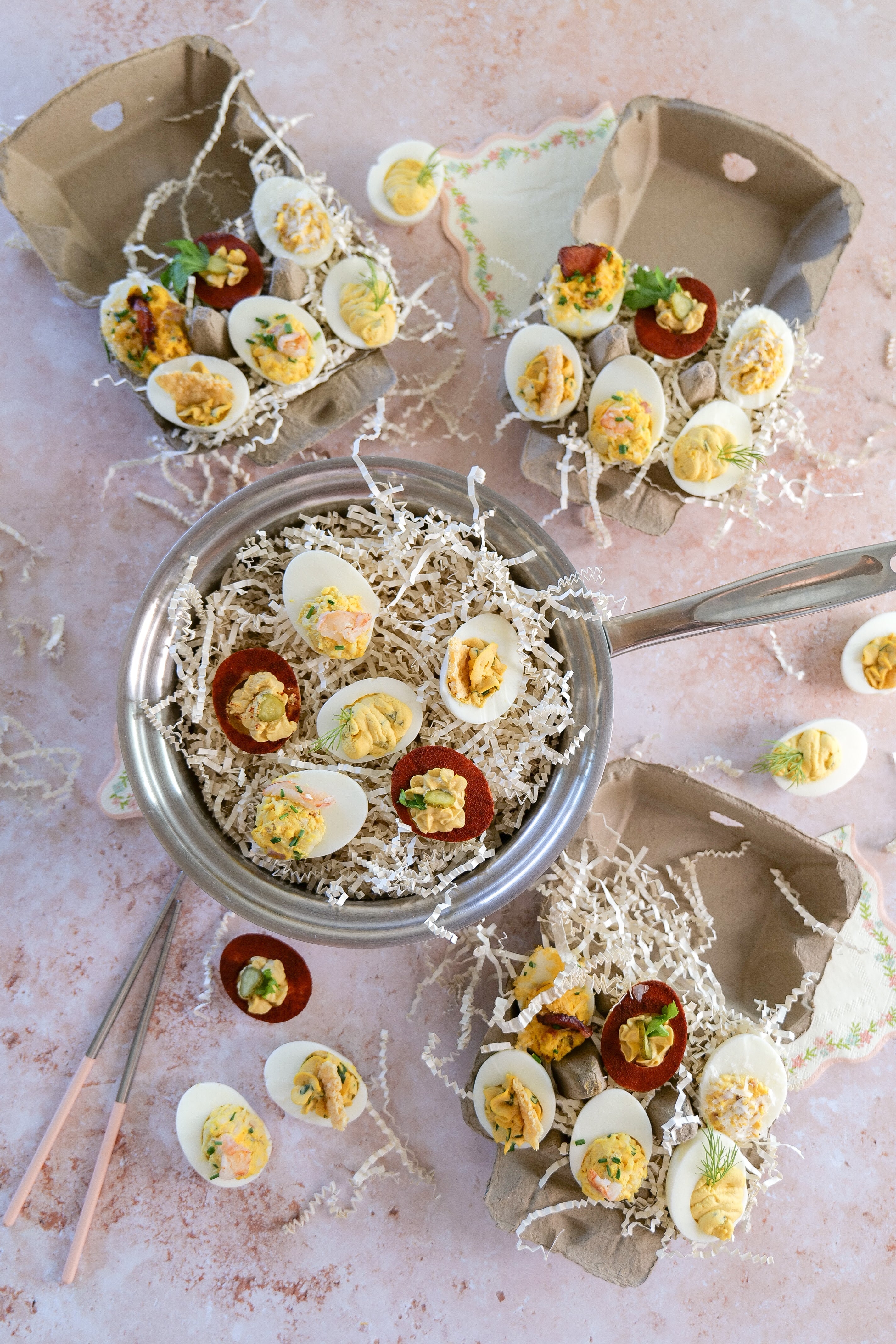Deviled eggs arranged in a stainless steel pot and recycled egg cartons on a pink marble surface with shredded paper