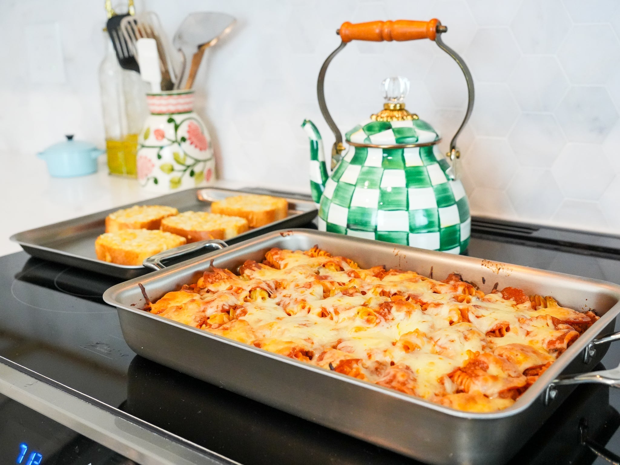 Pepperoni pasta bake in a stainless steel baking pan on a stovetop with garlic toast and a green checkered kettle