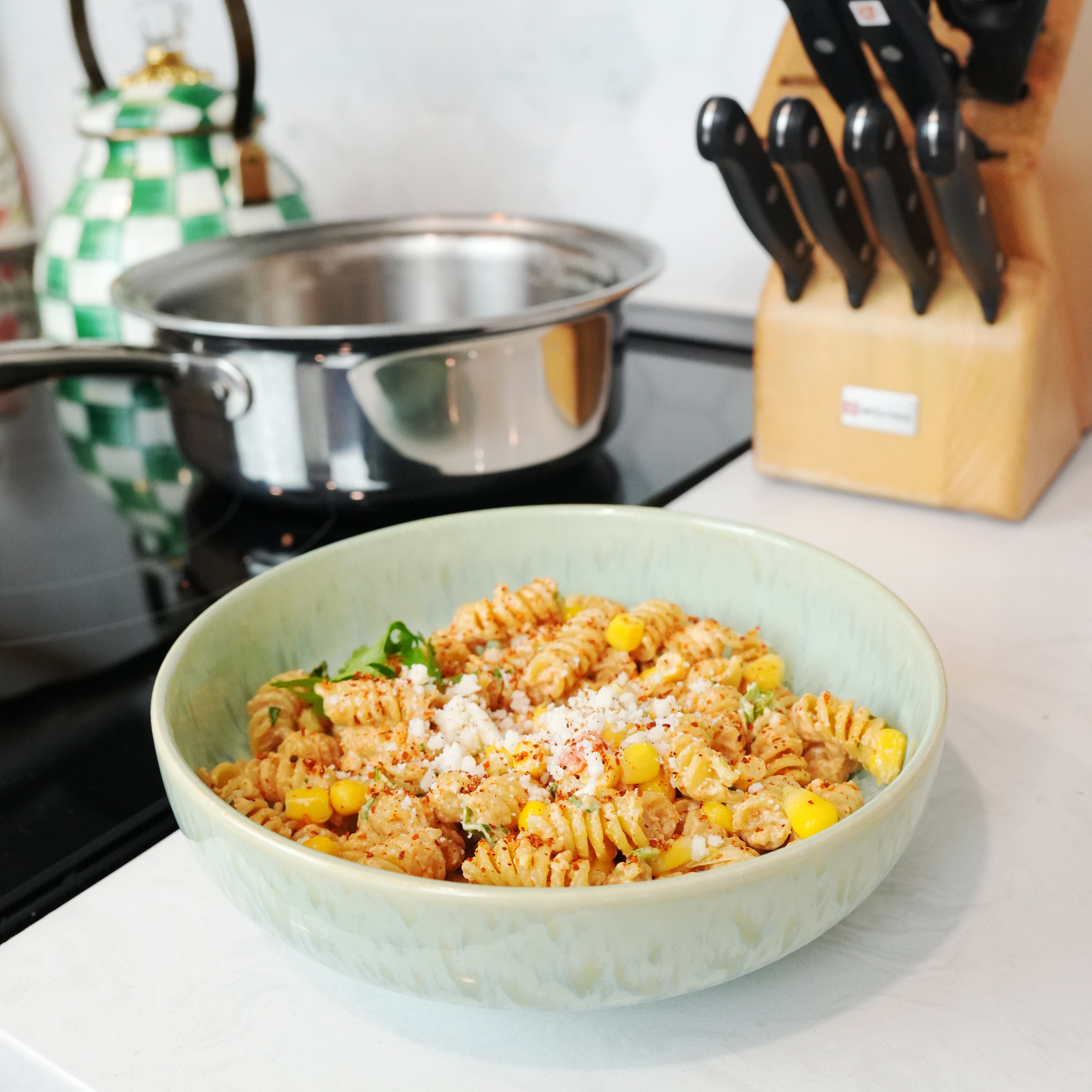 Close-up bowl of elote pasta salad on a kitchen counter with stainless steel stock pot and knife block behind