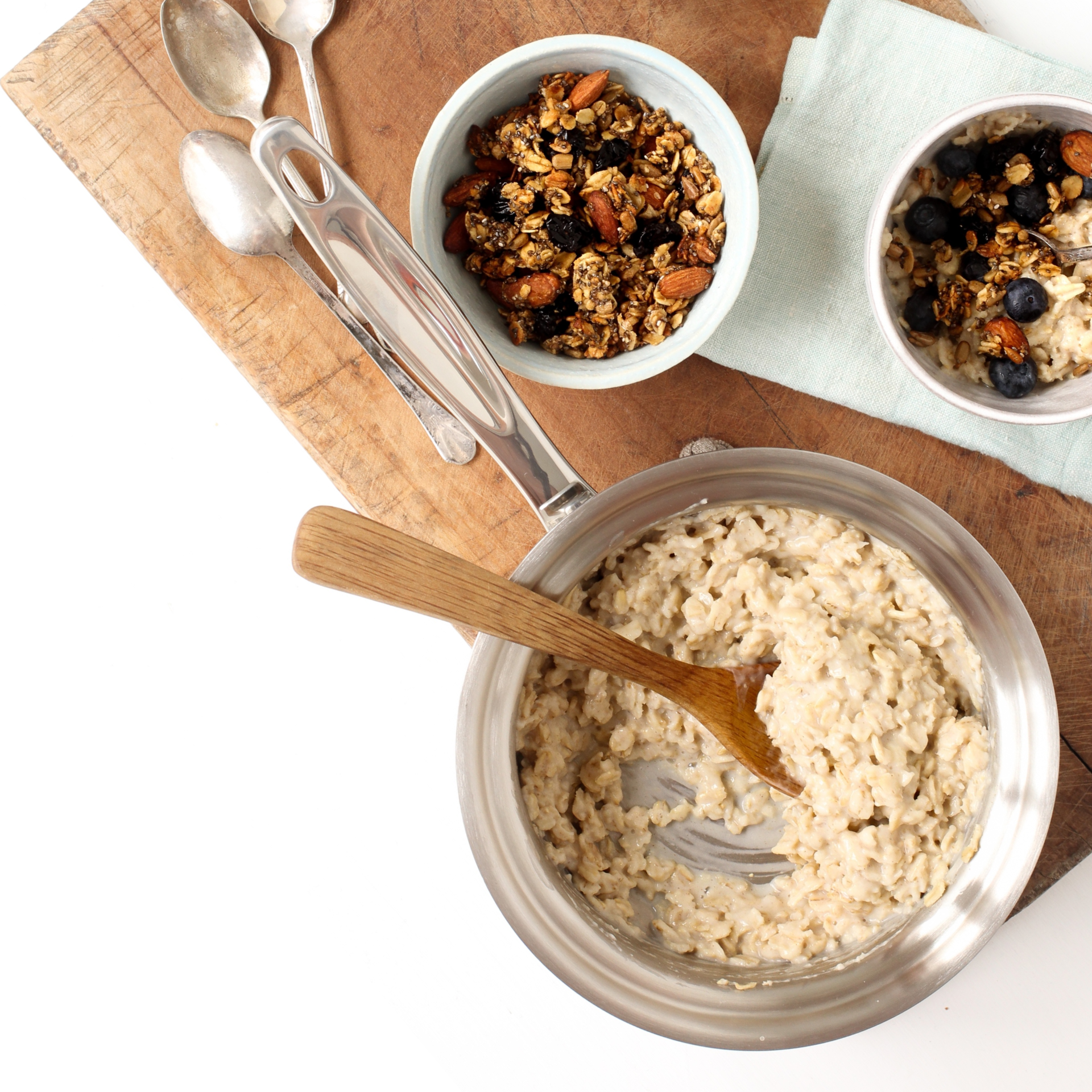 Cooked oatmeal in a 1.75 qt stainless steel saucepan with a wooden spoon, surrounded by bowls of granola and fruit.