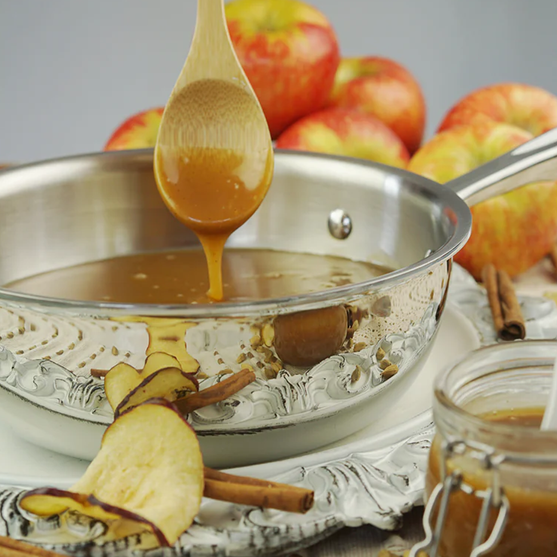 A stainless steel saucier with caramel sauce, a wooden spoon, apples, and cinnamon sticks.