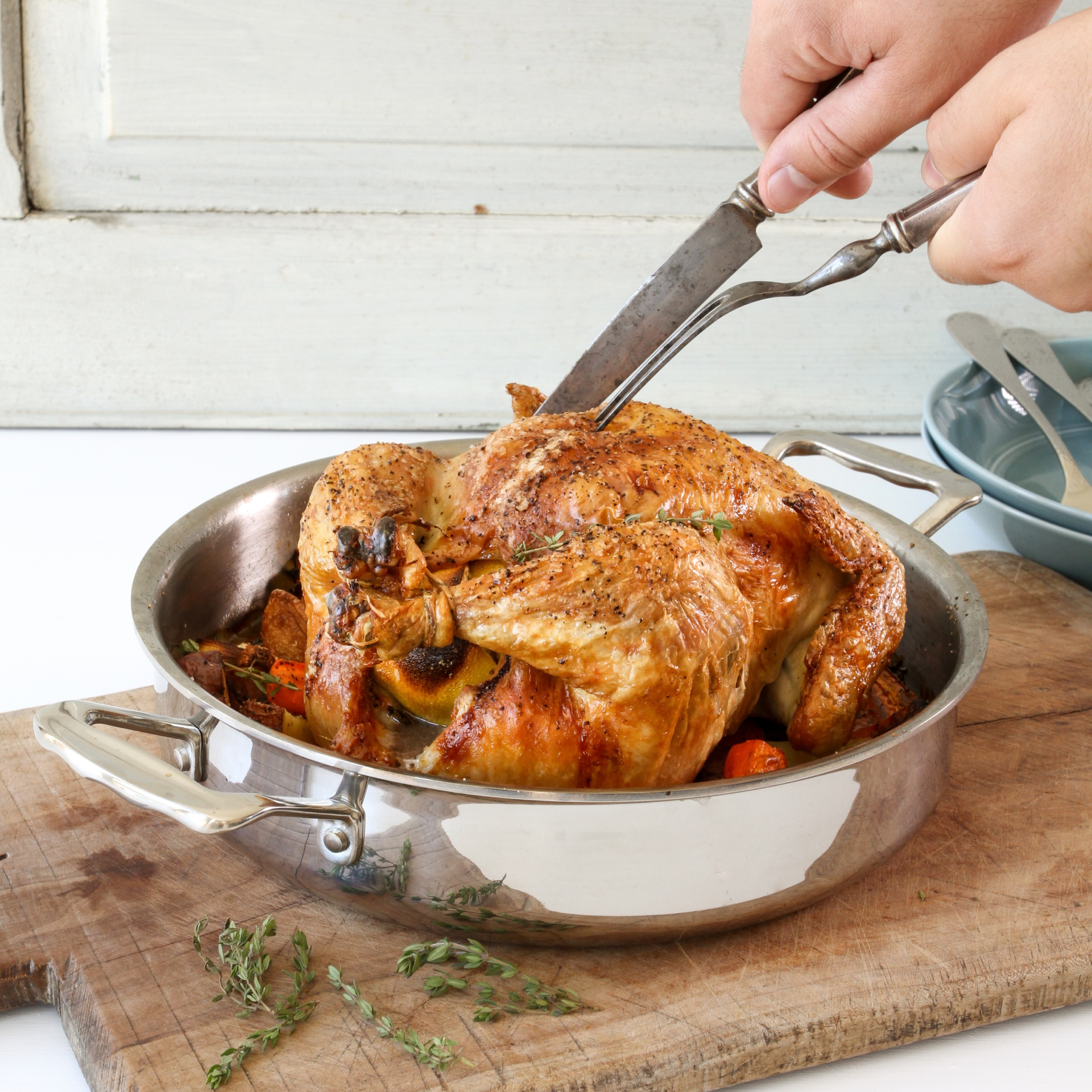 A hand carving a roasted chicken in a stainless steel roasting pan with vegetables and herbs.