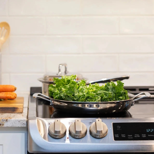A stainless steel fry pan on a stove with kale and carrots nearby.