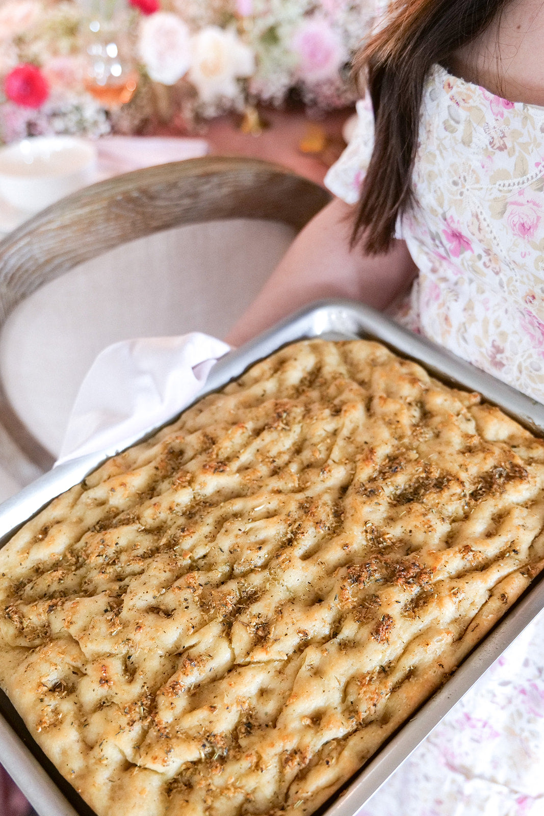 A person holding a tray of baked focaccia bread with herbs.