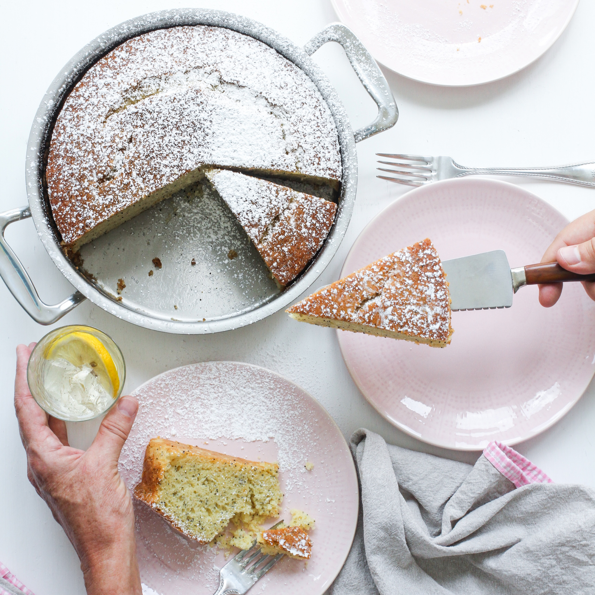 A hand serving a slice of cake from a round cake pan, with plates and a drink nearby.