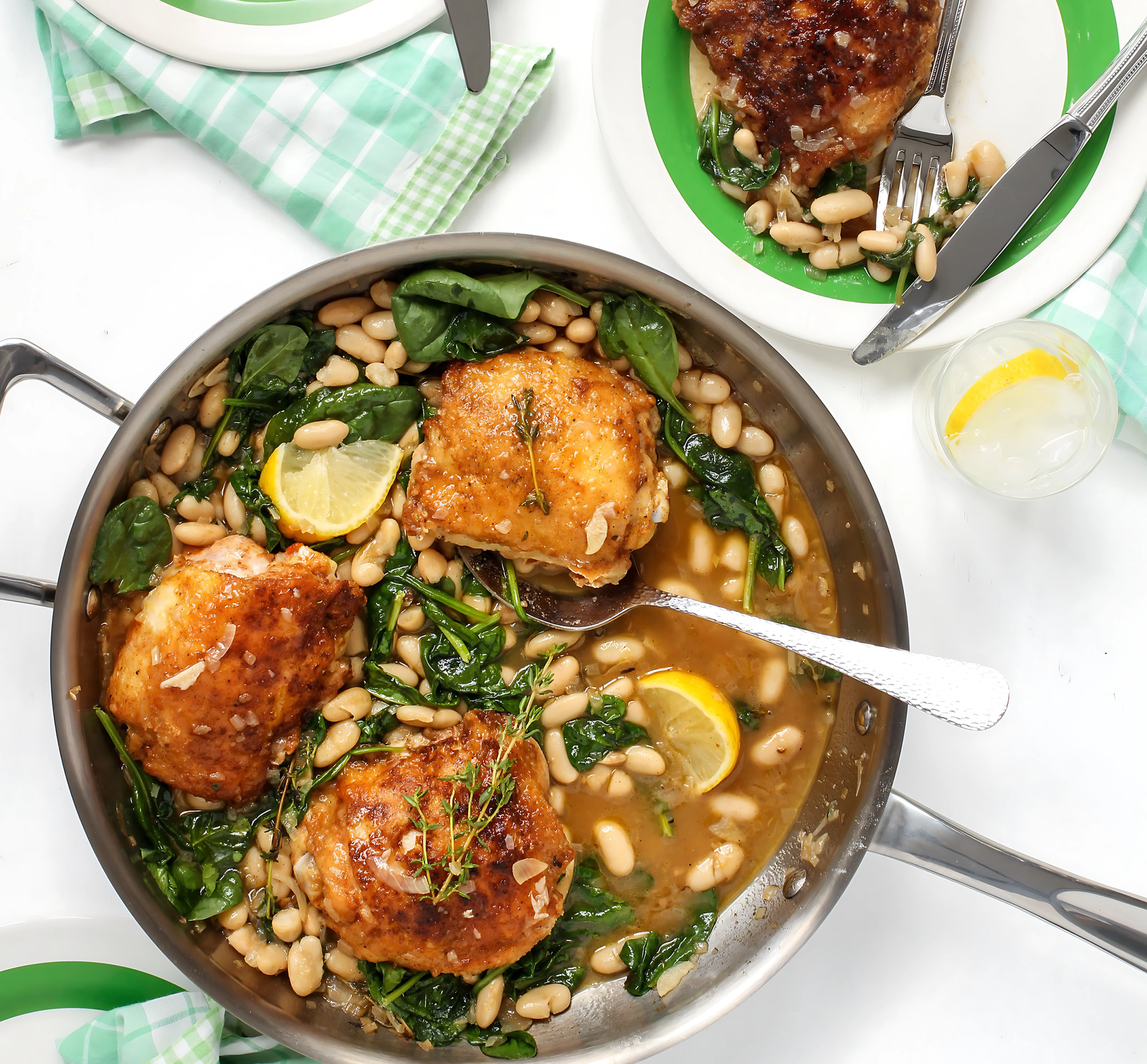 Skillet with chicken, spinach, and white beans on a white table with plates and napkins.