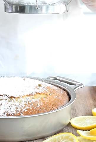 Roasting pan holding a baked lemon cake being dusted with powdered sugar on a wooden surface with lemon slices