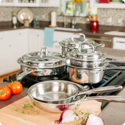 Stainless steel cookware set on a kitchen counter with tomatoes and herbs.