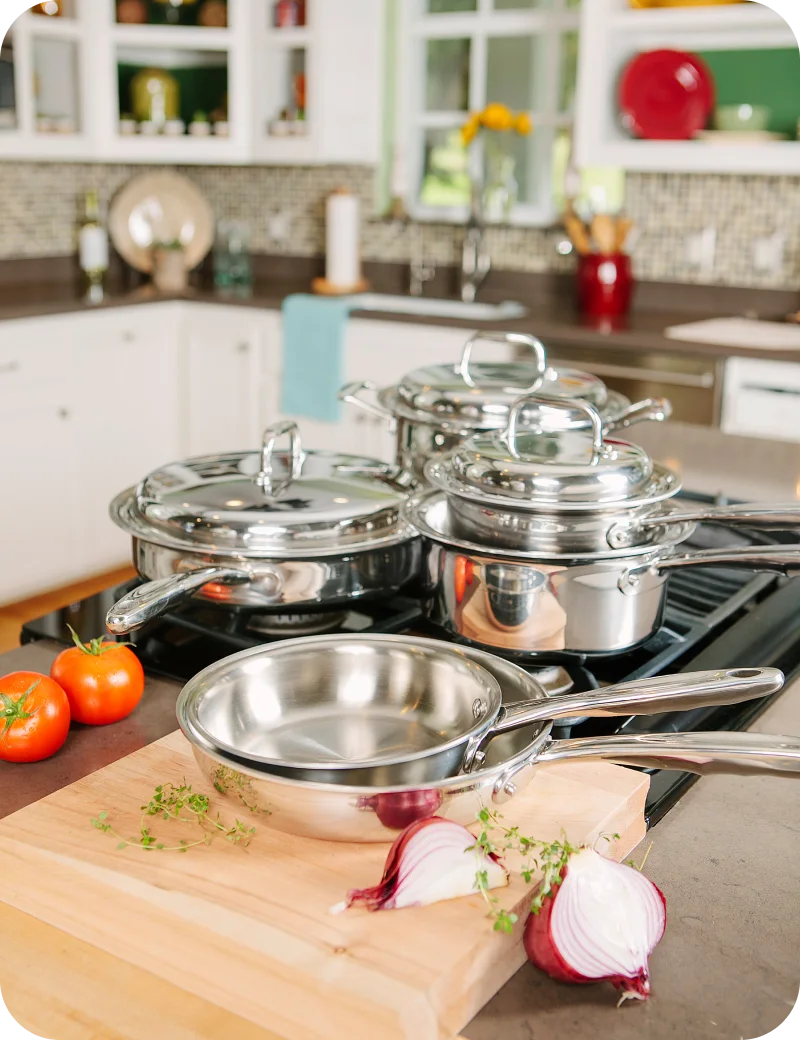 Set of stainless steel cookware on a kitchen stove with vegetables on a cutting board.
