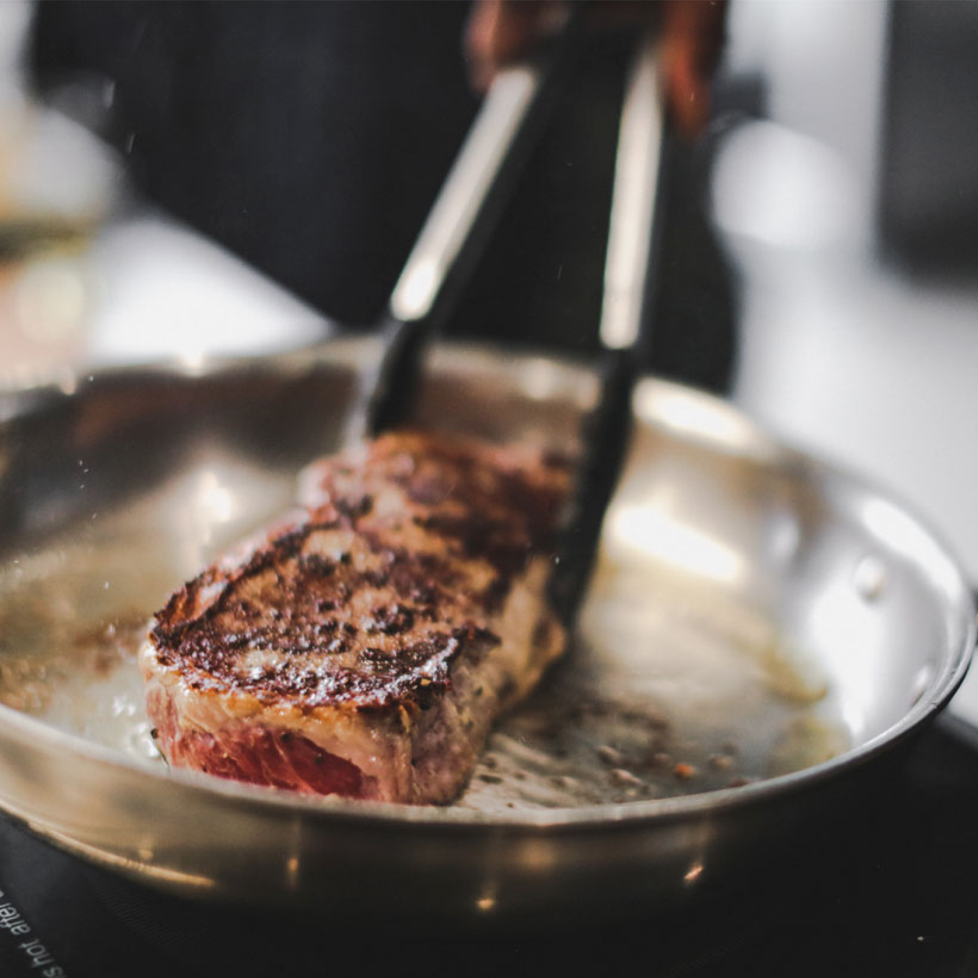 A steak being cooked in a stainless steel fry pan with short handles.