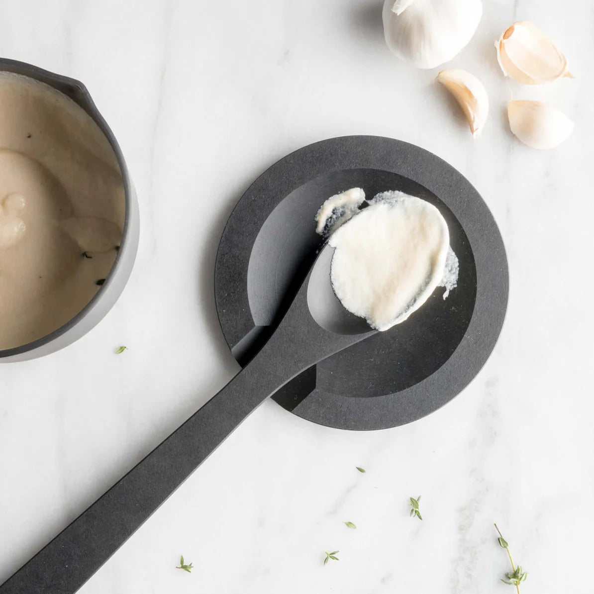 A slate round utensil rest with a spoon holding a creamy substance, surrounded by garlic cloves and herbs.