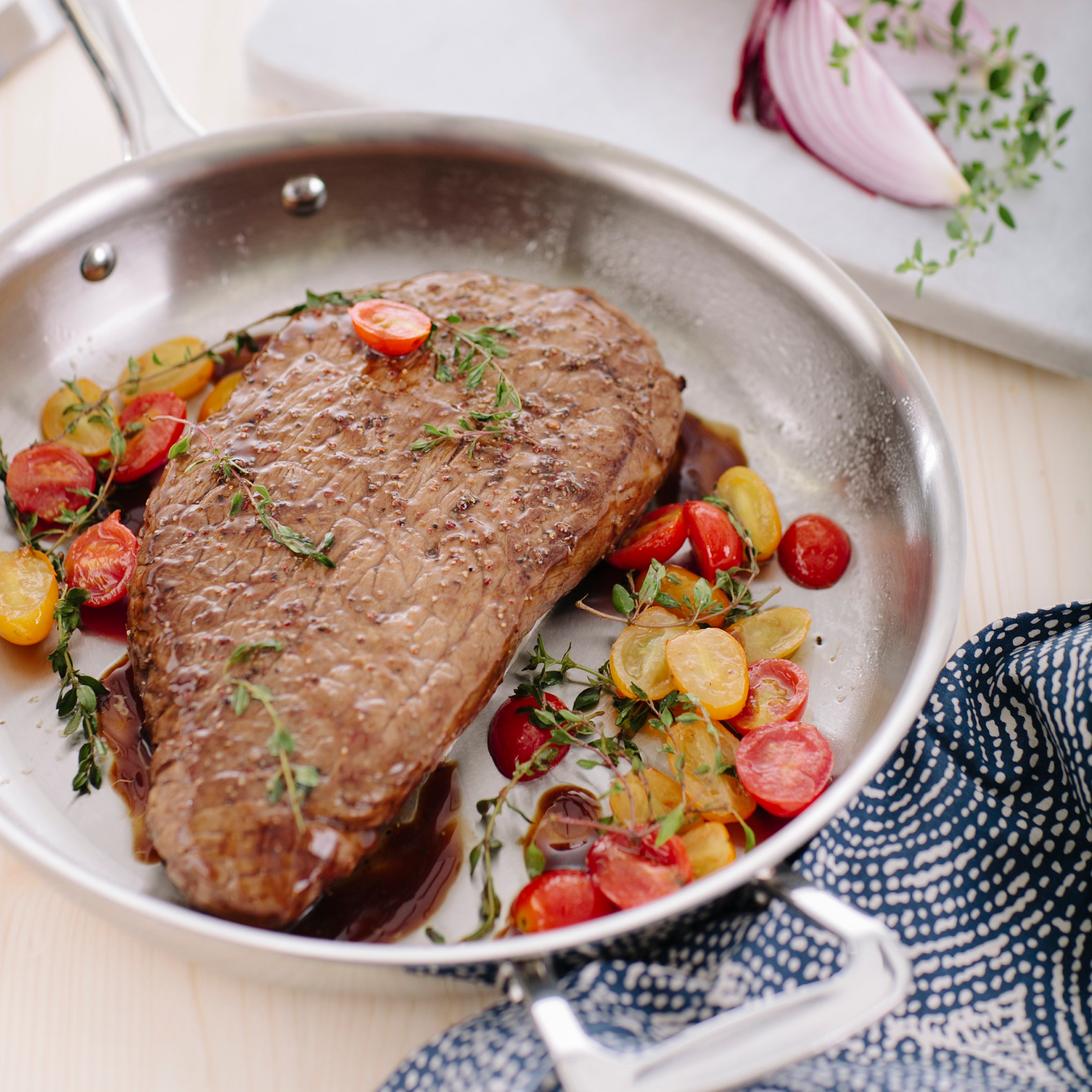 Steak with herbs and tomatoes in a stainless steel pan on a table.