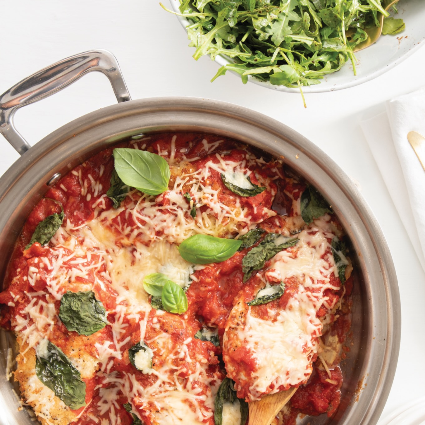 A sauté pan filled with chicken, marinara sauce, cheese, and fresh basil, with a side salad in a bowl.