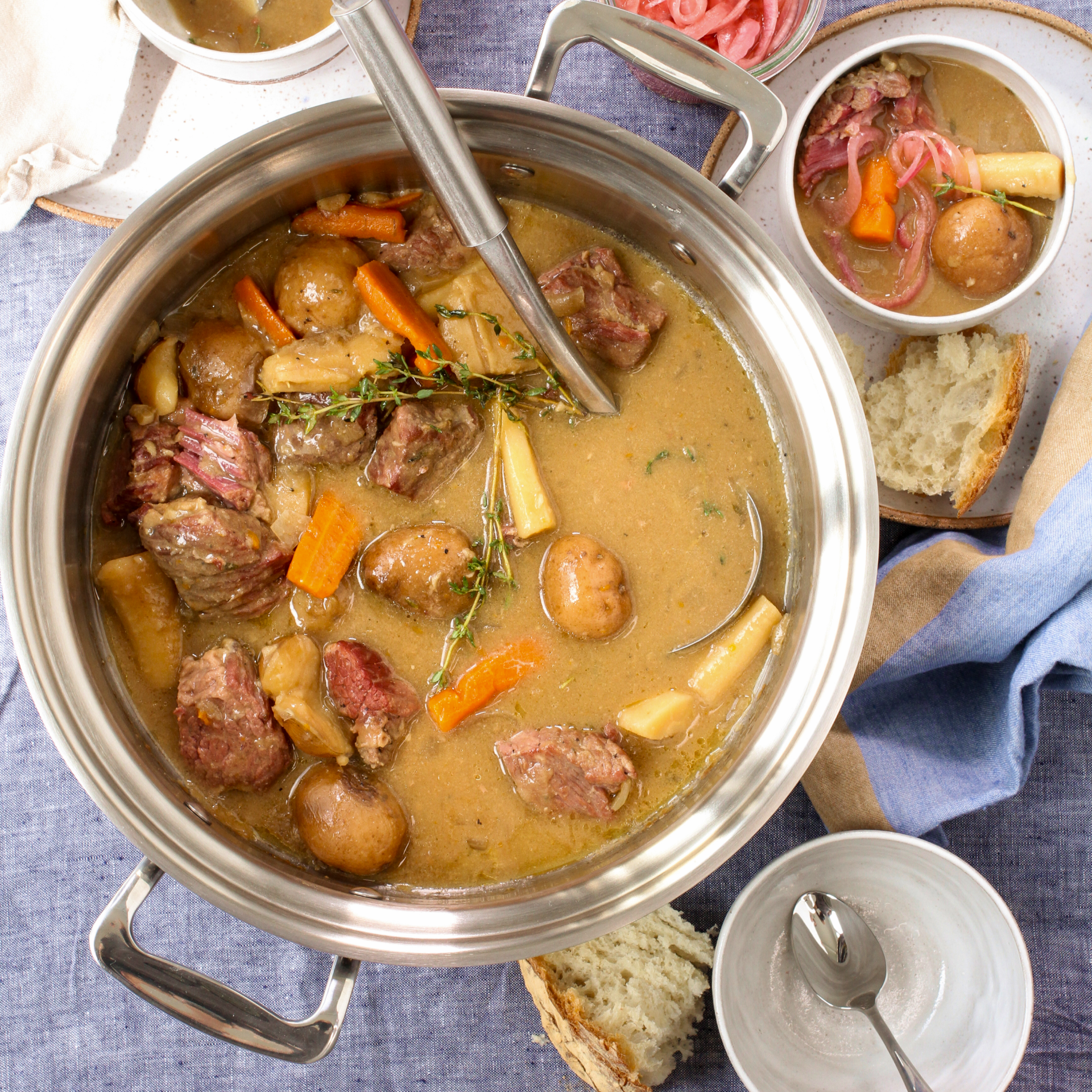 A 6-quart stainless steel stock pot filled with beef stew, carrots, potatoes, and herbs on a blue tablecloth.