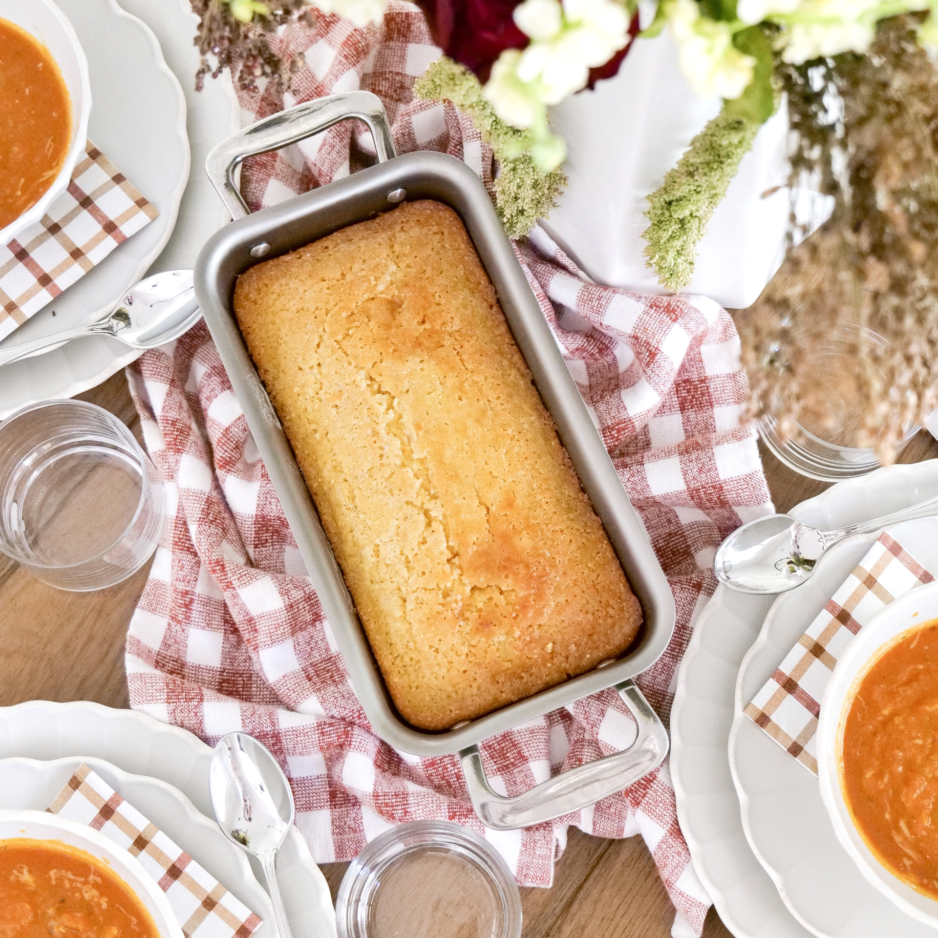 A freshly baked loaf of bread in a stainless steel loaf pan on a table with soup and utensils.