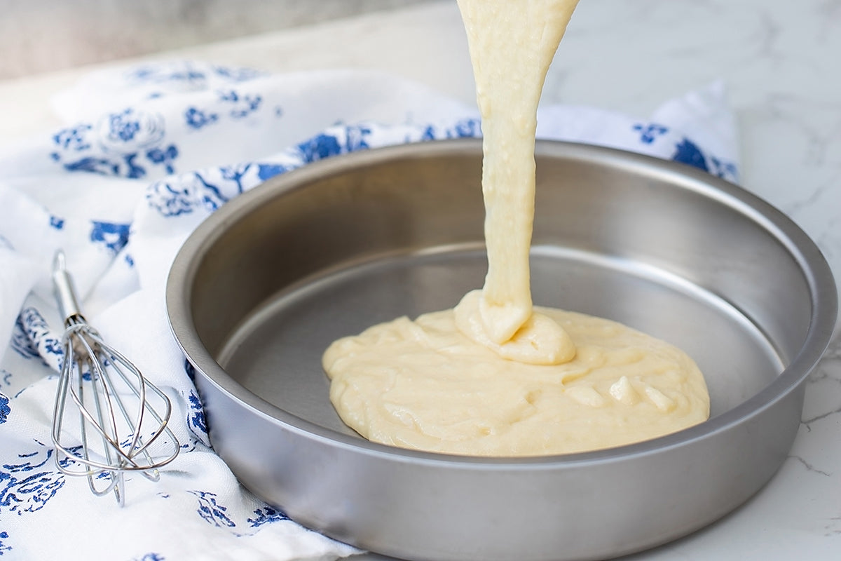 Batter being poured into a 9" round cake pan on a marble surface with a whisk nearby.
