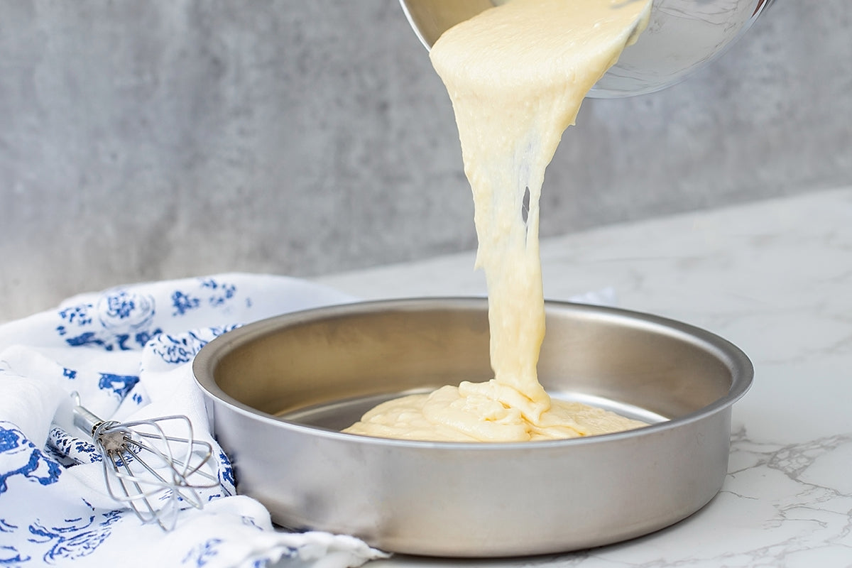 Batter being poured into a 9" round stainless steel cake pan on a marble surface.