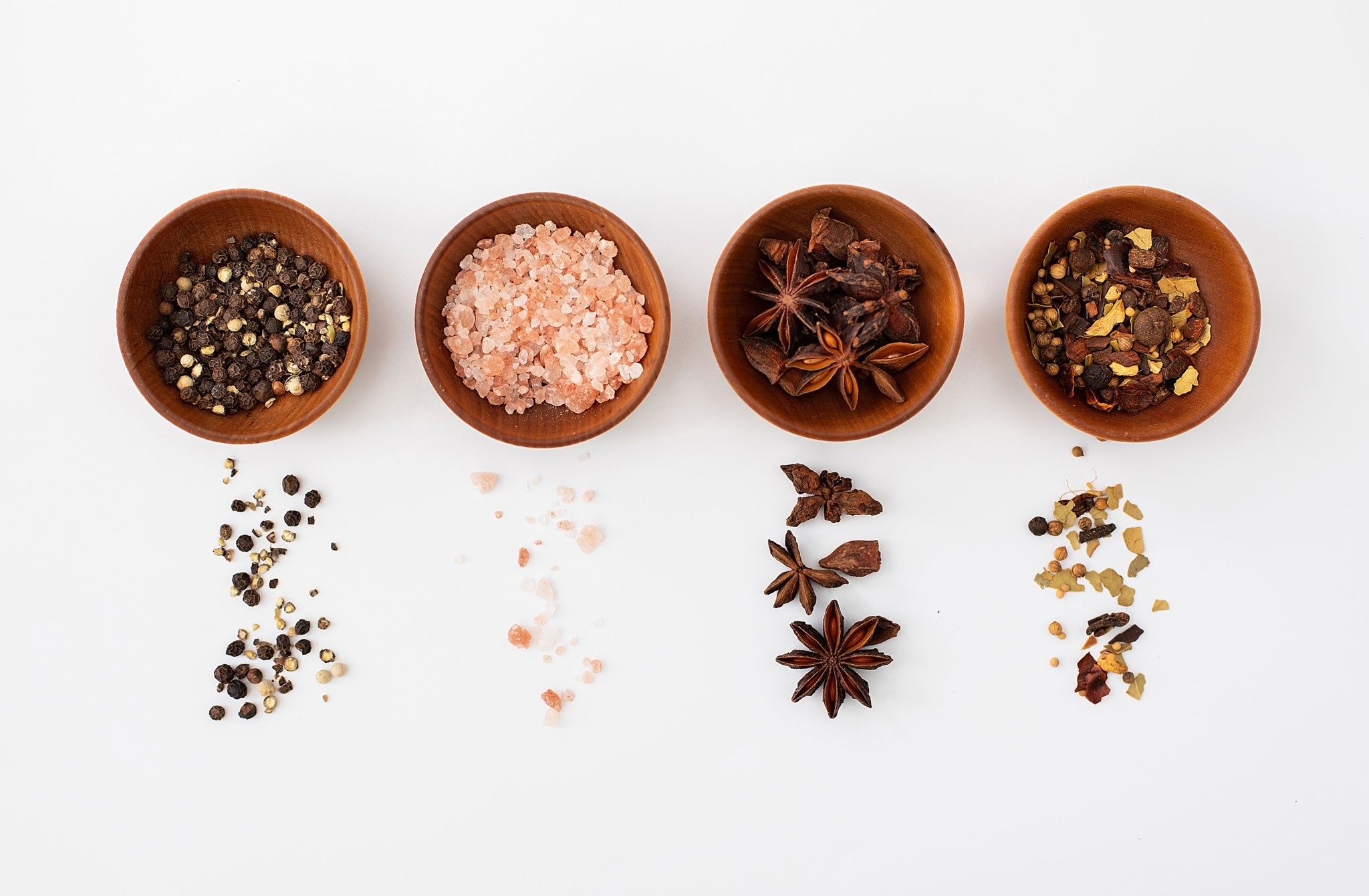 Four wooden condiment cups filled with various spices including pink salt, peppercorns, and star anise on a white background.