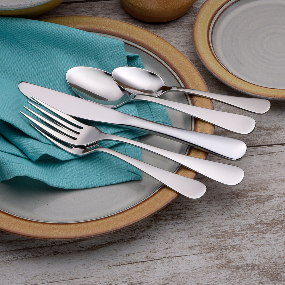 Five pieces of Annapolis flatware including a knife, fork, and spoons on a teal napkin and plates.
