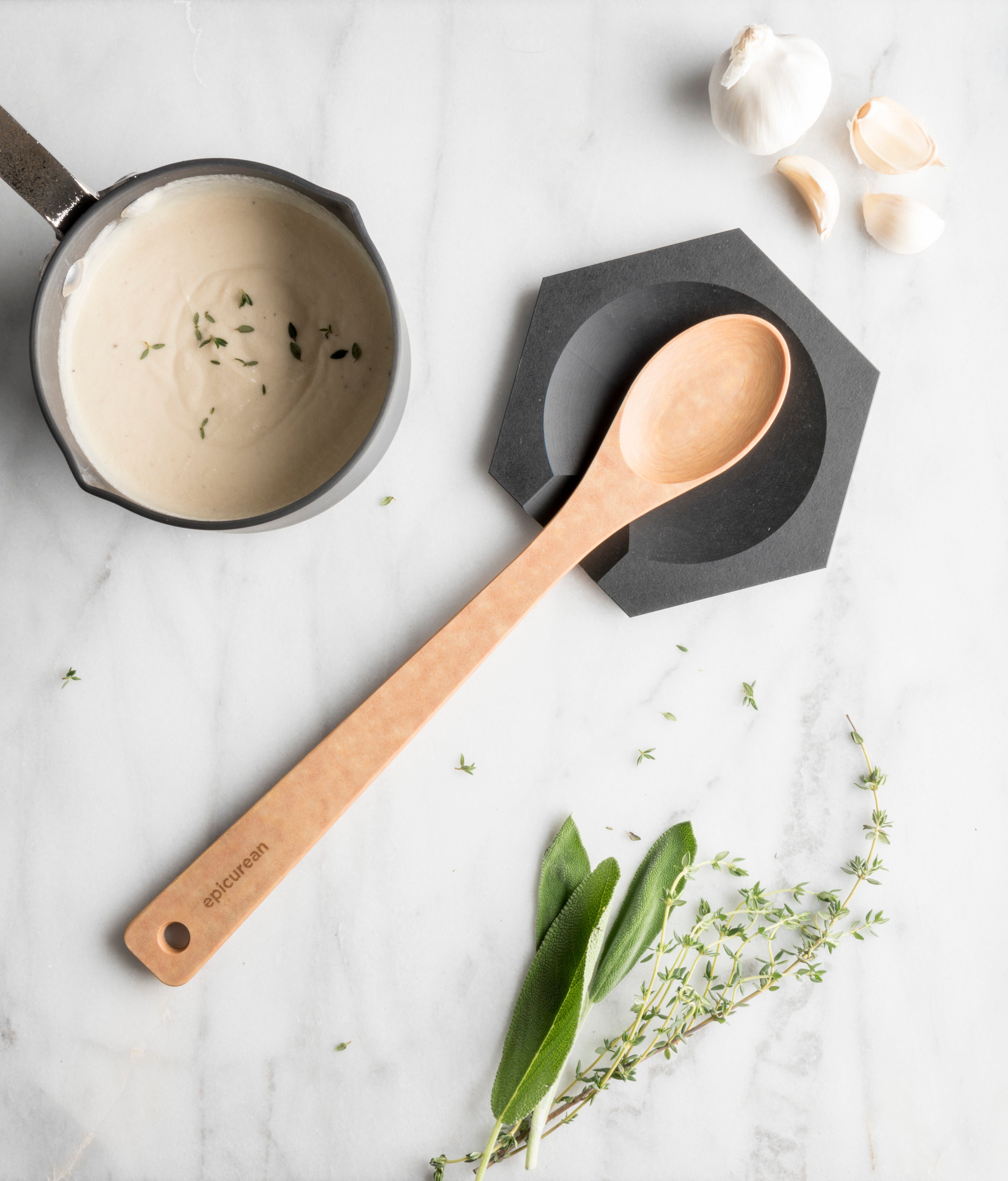 A wooden spoon on a slate hex tool rest with a pot of sauce and herbs.