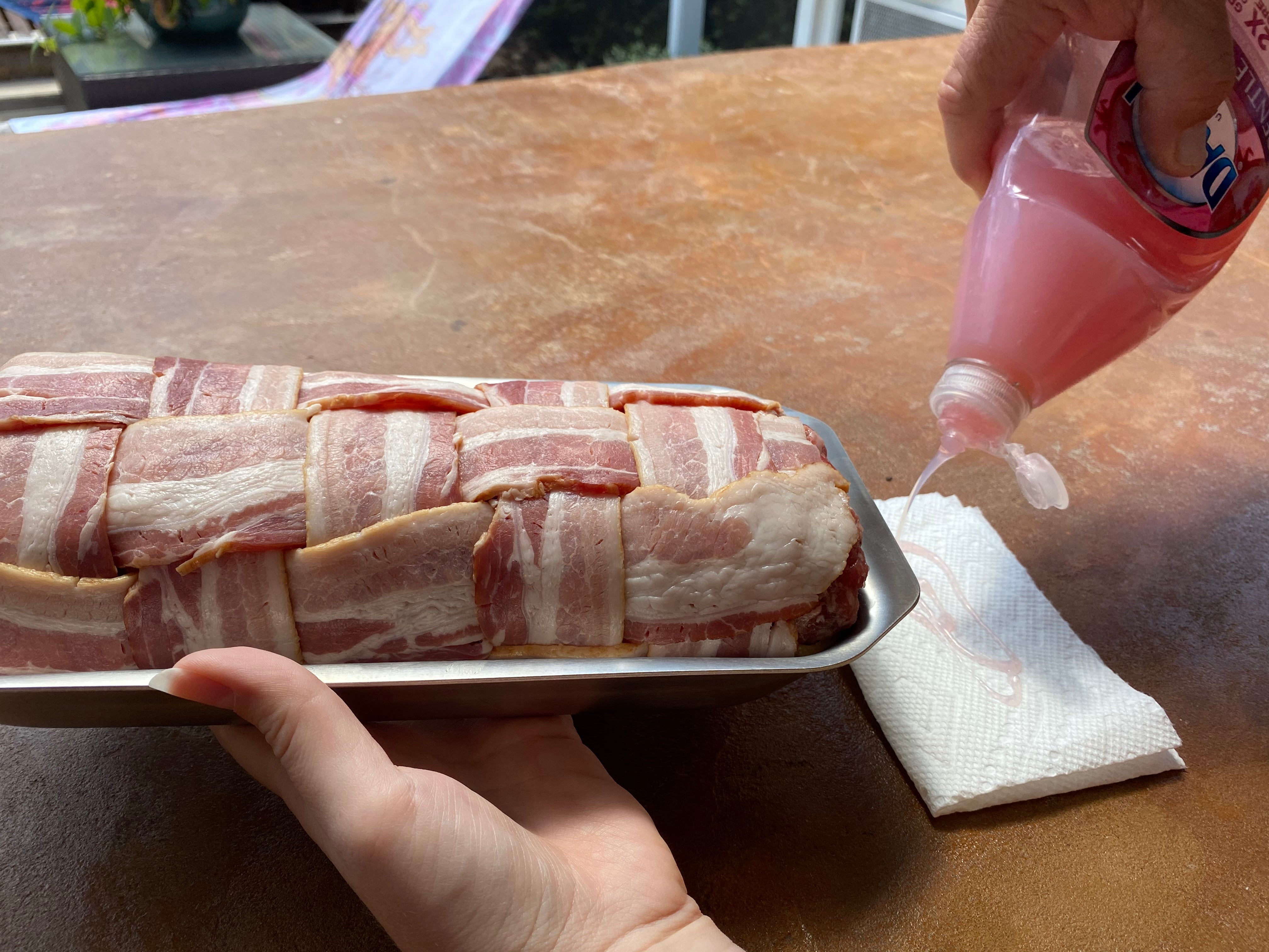 A hand holding a tray of bacon-wrapped meat with a bottle of sauce pouring over it.