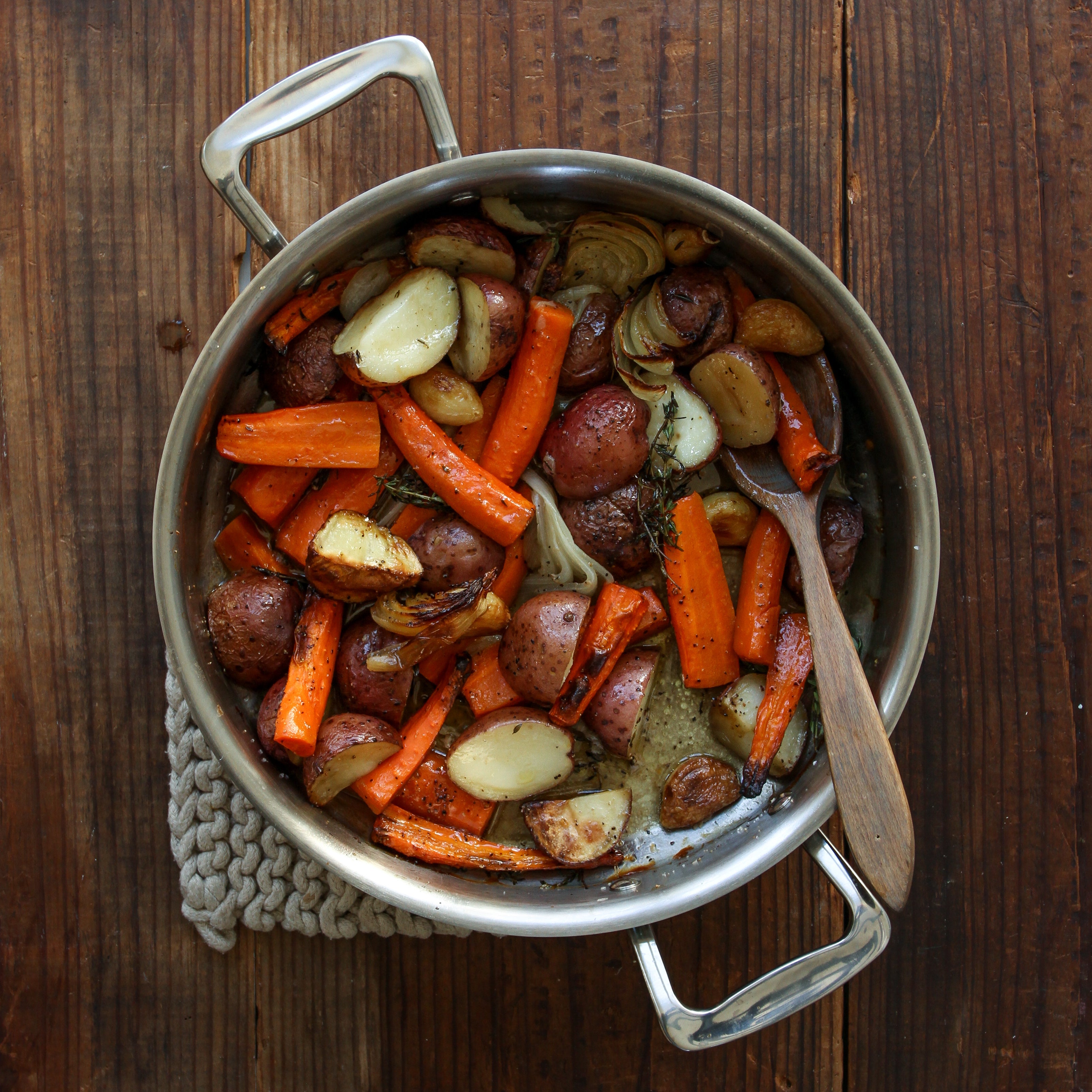 A stainless steel roasting pan filled with roasted vegetables including carrots, potatoes, and onions.
