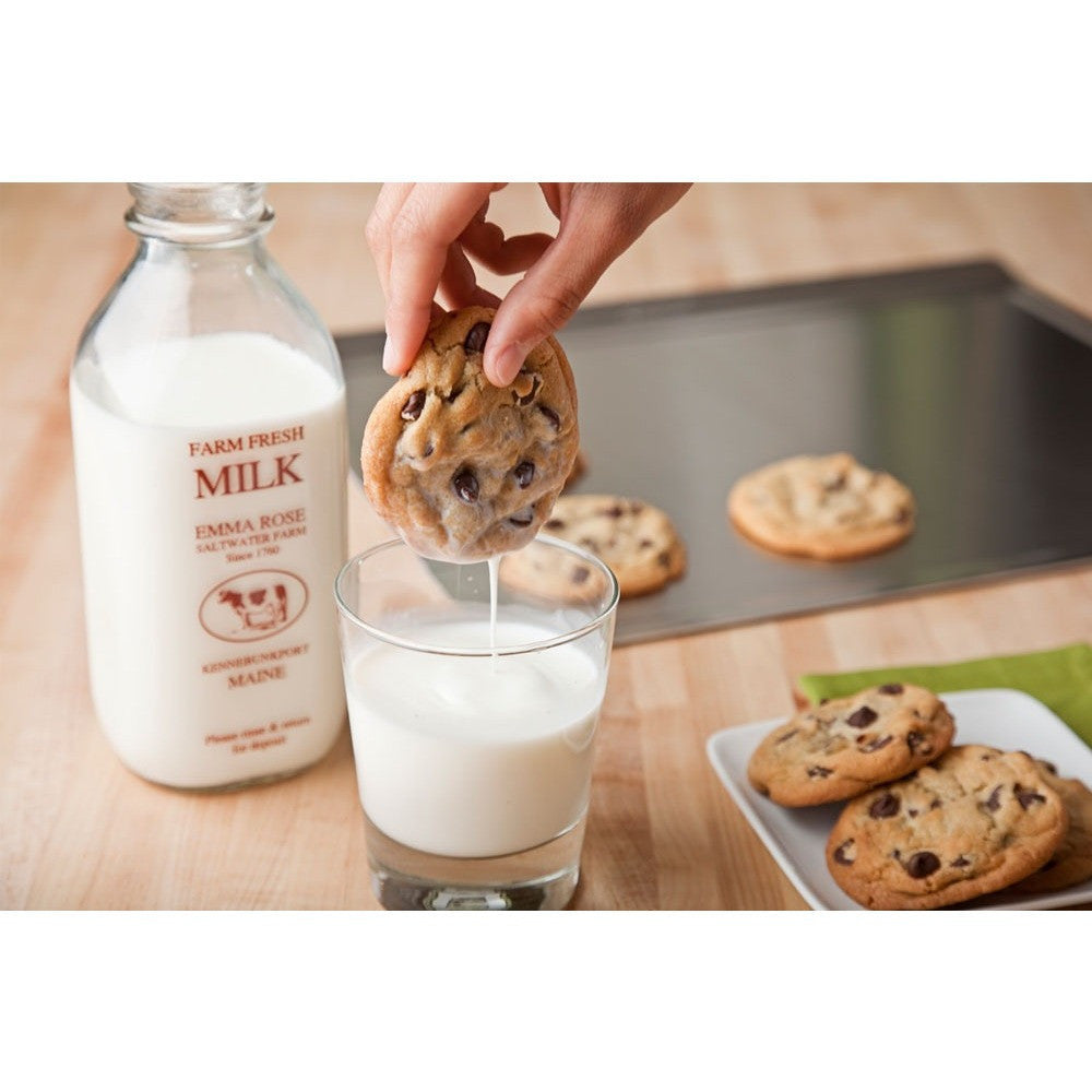 A hand dipping a chocolate chip cookie into a glass of milk, with cookies on a tray in the background.