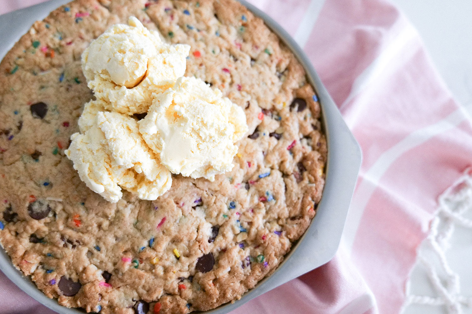 A round cookie cake topped with scoops of vanilla ice cream in a baking pan