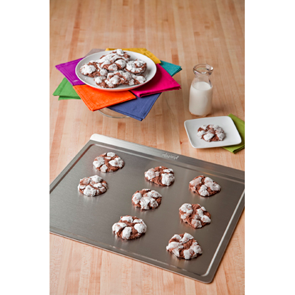 A large stainless steel cookie sheet with cookies on it, surrounded by colorful napkins and a glass of milk.