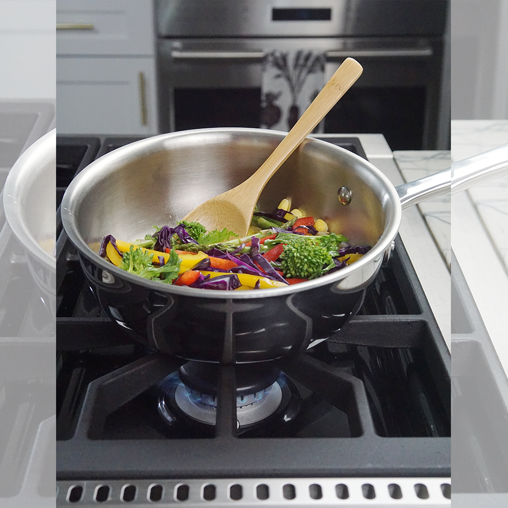 A 2-quart stainless steel saucier on a stove with vegetables being sautéed and a wooden spoon.